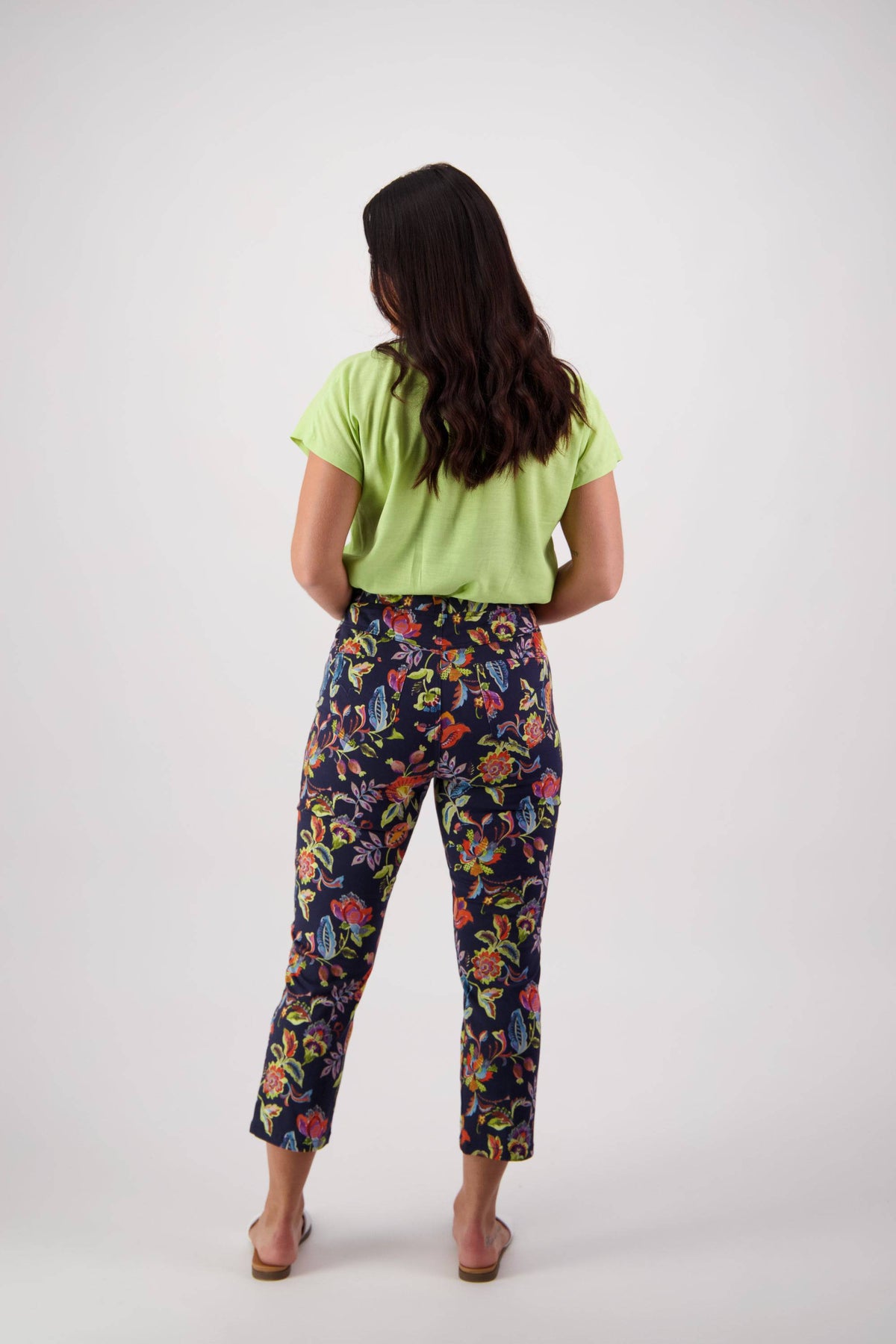 A woman with wavy dark hair faces away, wearing a light green top and Vassalli's Sierra 7/8 Pant - Brazil, featuring a colorful floral pattern and slim leg pull-on design. The background is plain and light colored.