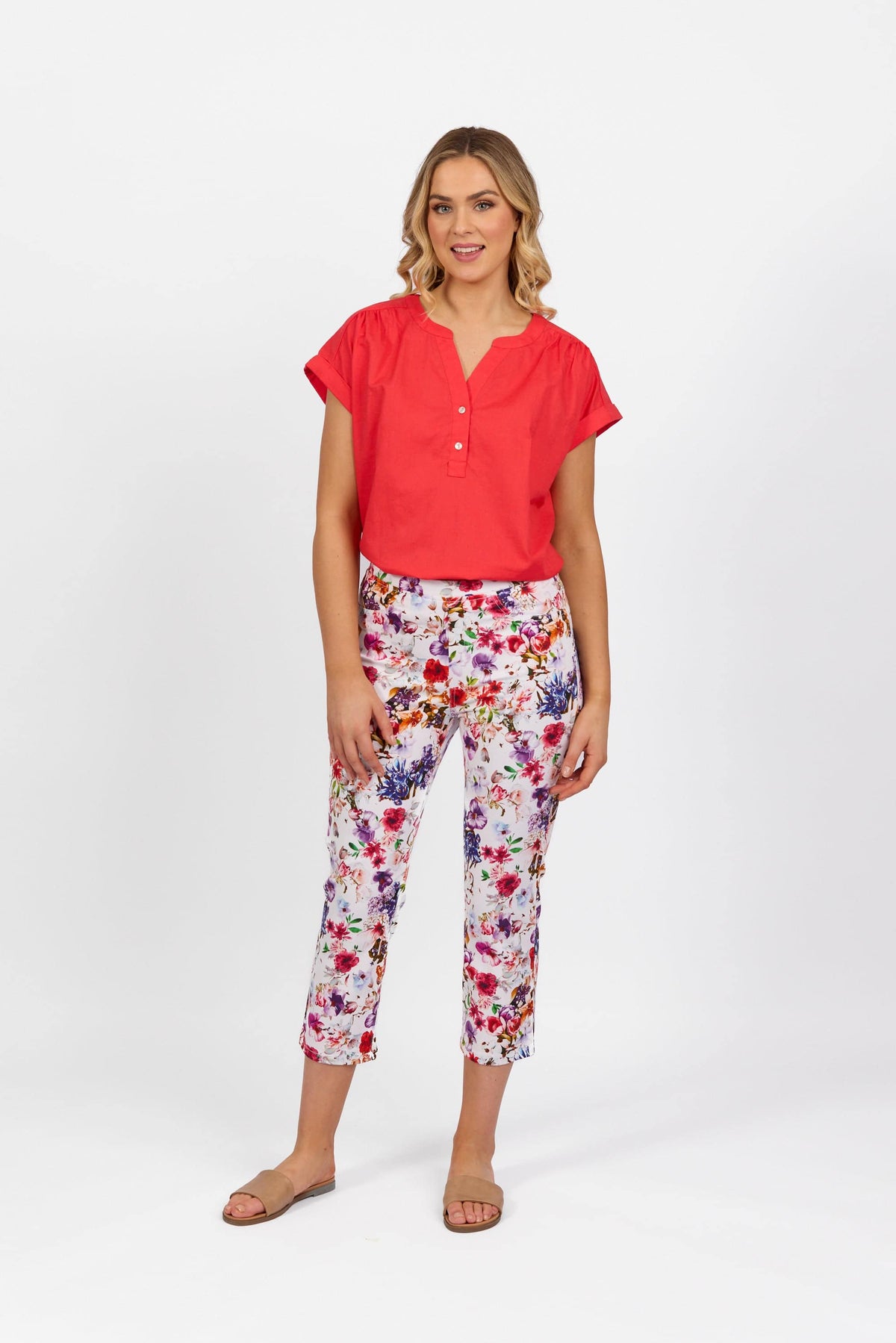 A woman with shoulder-length blonde hair smiles against a white background, wearing a bright red short-sleeve blouse, Vassalli Sierra 7/8 Pant - Burst, and beige flat sandals.
