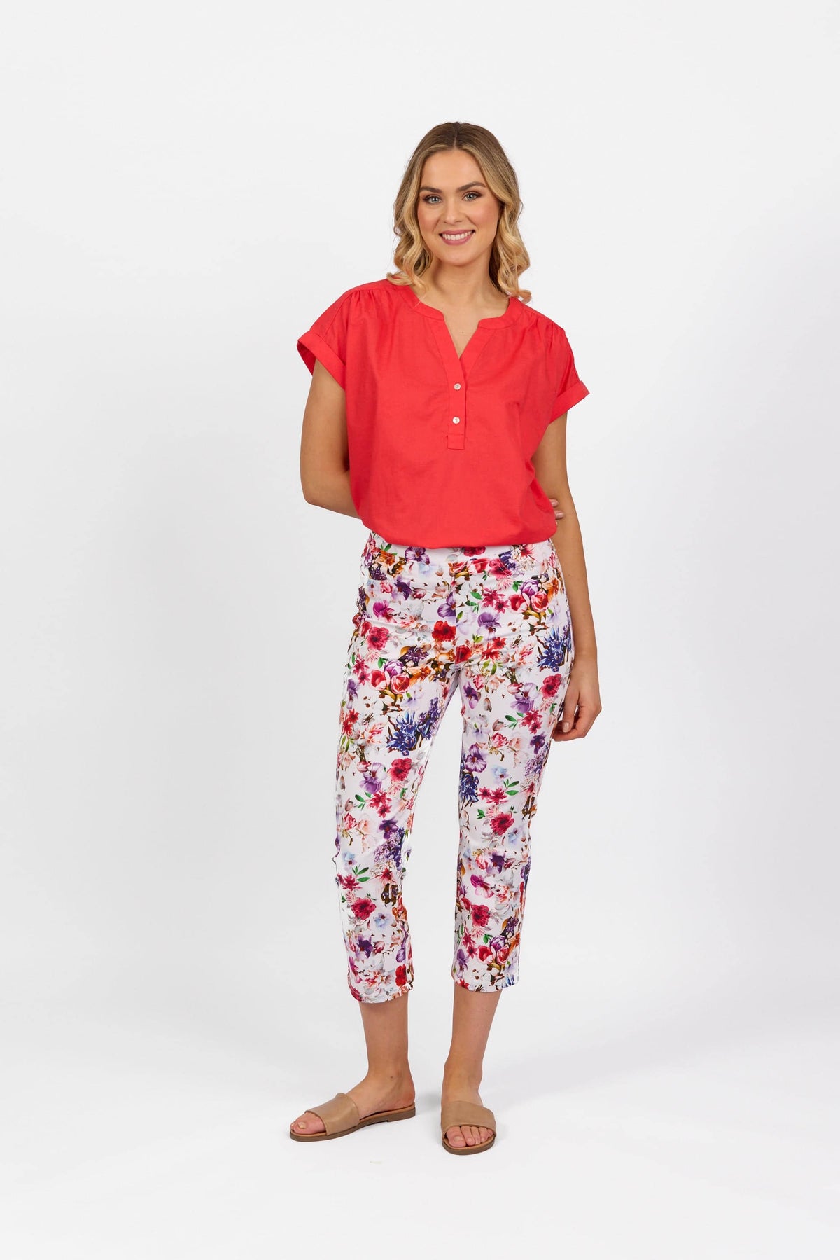 A woman smiles and poses against a white background, wearing a short-sleeve red blouse, Vassalli Sierra 7/8 Pant - Burst, and tan sandals.