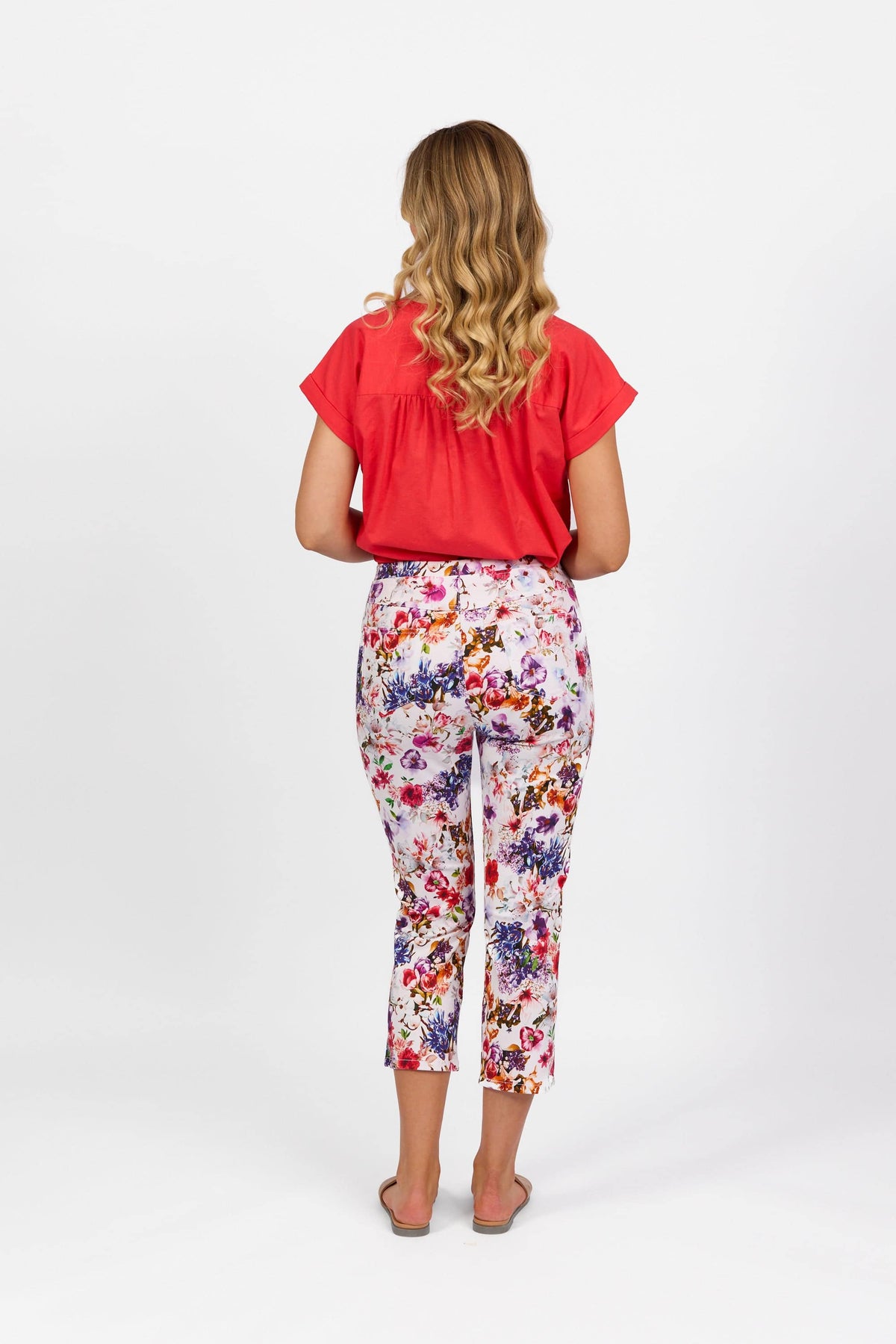 A woman with long, wavy blonde hair stands facing away against a plain white background, wearing a red short-sleeve top, Vassalli Sierra 7/8 Pant - Burst, and sandals.