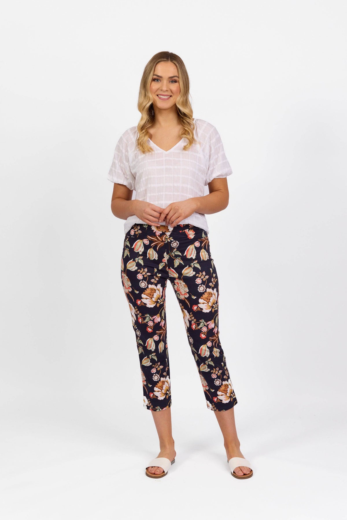 A woman with loose waves smiles against a white background, wearing a white short-sleeve top, Vassalli’s Sierra 7/8 Pant - Freya in a printed slim leg stretch fabric, and white slide sandals.