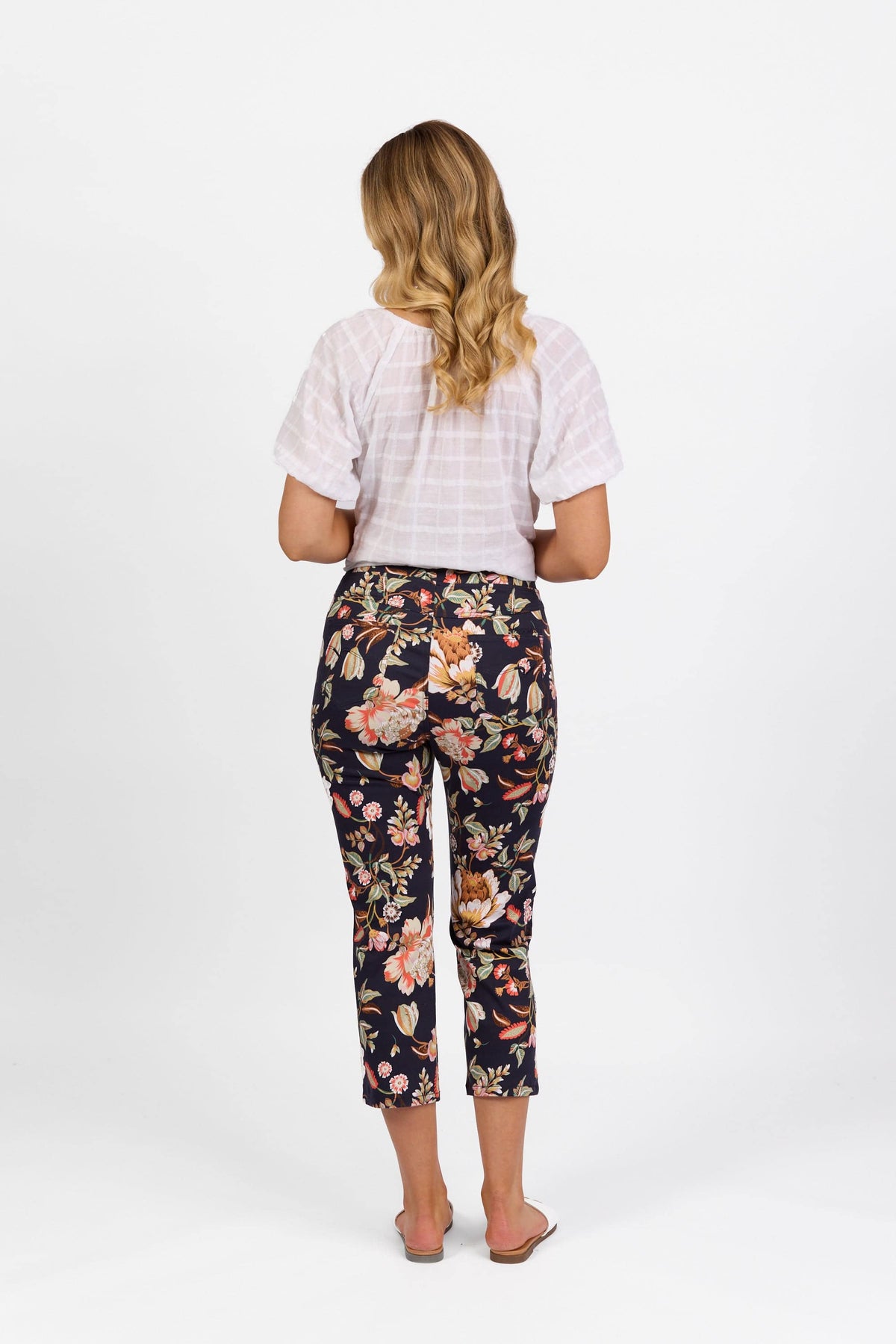 A woman with wavy blonde hair faces away, wearing a short-sleeve white blouse and Vassalli's Sierra 7/8 Pant - Freya, printed slim-leg pull-on pants in lightweight stretch fabric. She is barefoot against a plain white background.