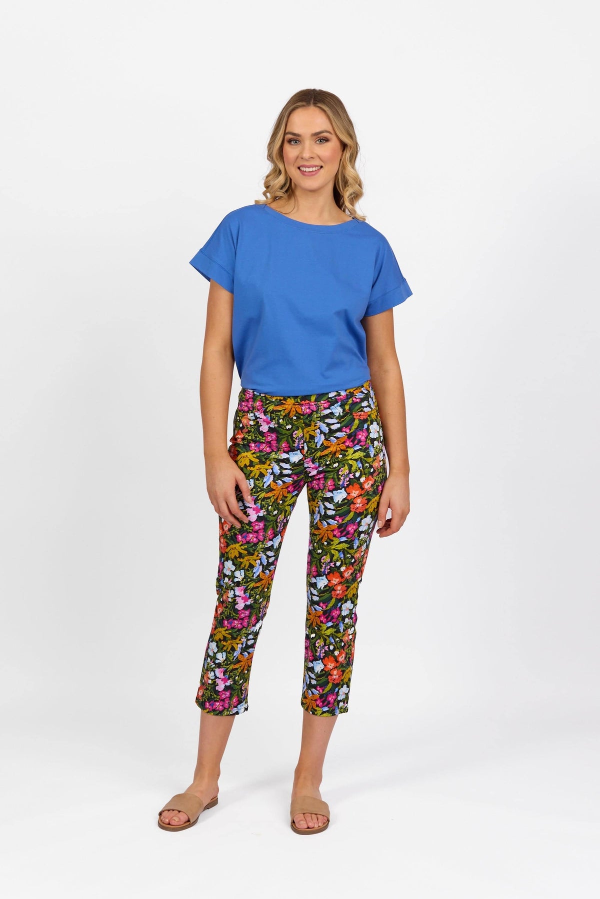 A woman smiles against a white background, wearing a bright blue short-sleeve top and Vassalli Sierra 7/8 Pant - Lola, pairing the floral cropped pants with beige sandals. Her shoulder-length hair is wavy.