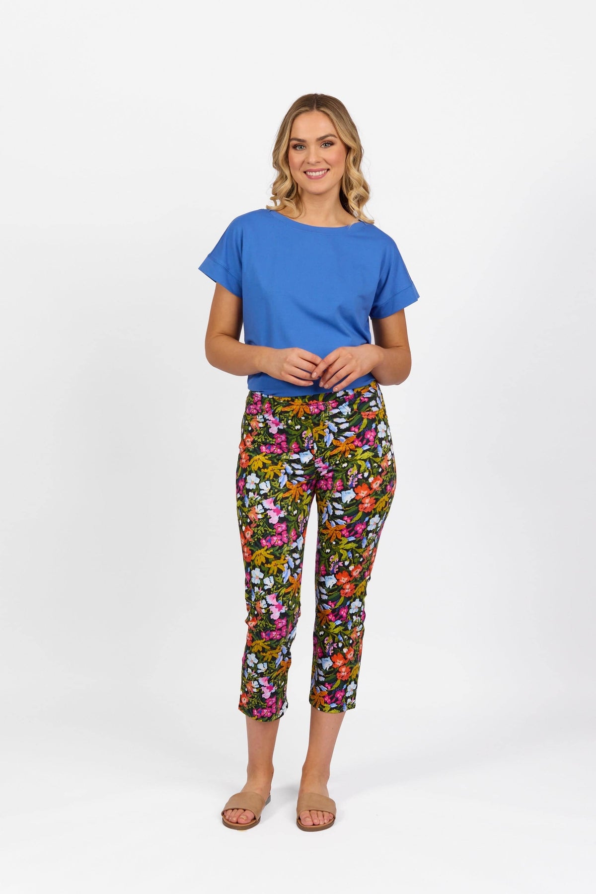 A woman stands smiling against a white background, wearing a blue short-sleeve top, Vassalli Sierra 7/8 Pant - Lola, and tan sandals.