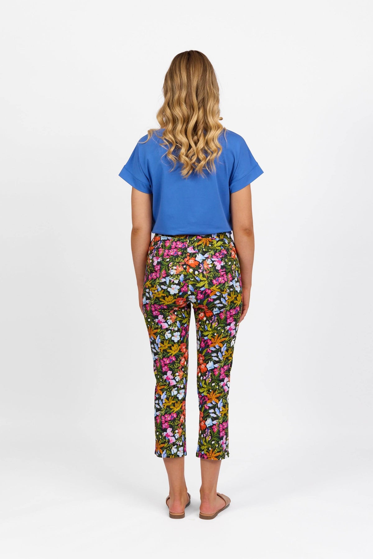 A woman with long wavy blonde hair stands facing away, wearing a blue short-sleeve top, Vassalli Sierra 7/8 Pant - Lola, and flat sandals against a white background.