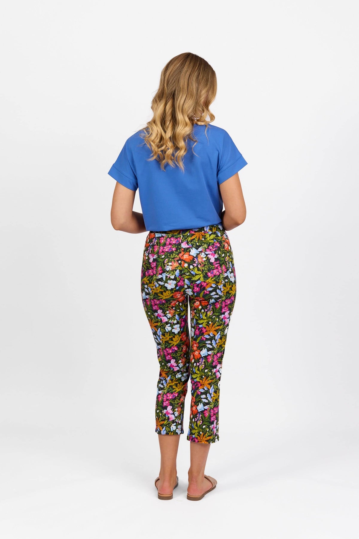 A woman with long blonde hair faces away, wearing a blue top and Vassalli Sierra 7/8 Pant - Lola, a lightweight pull-on floral pant, paired with sandals against a plain white background.