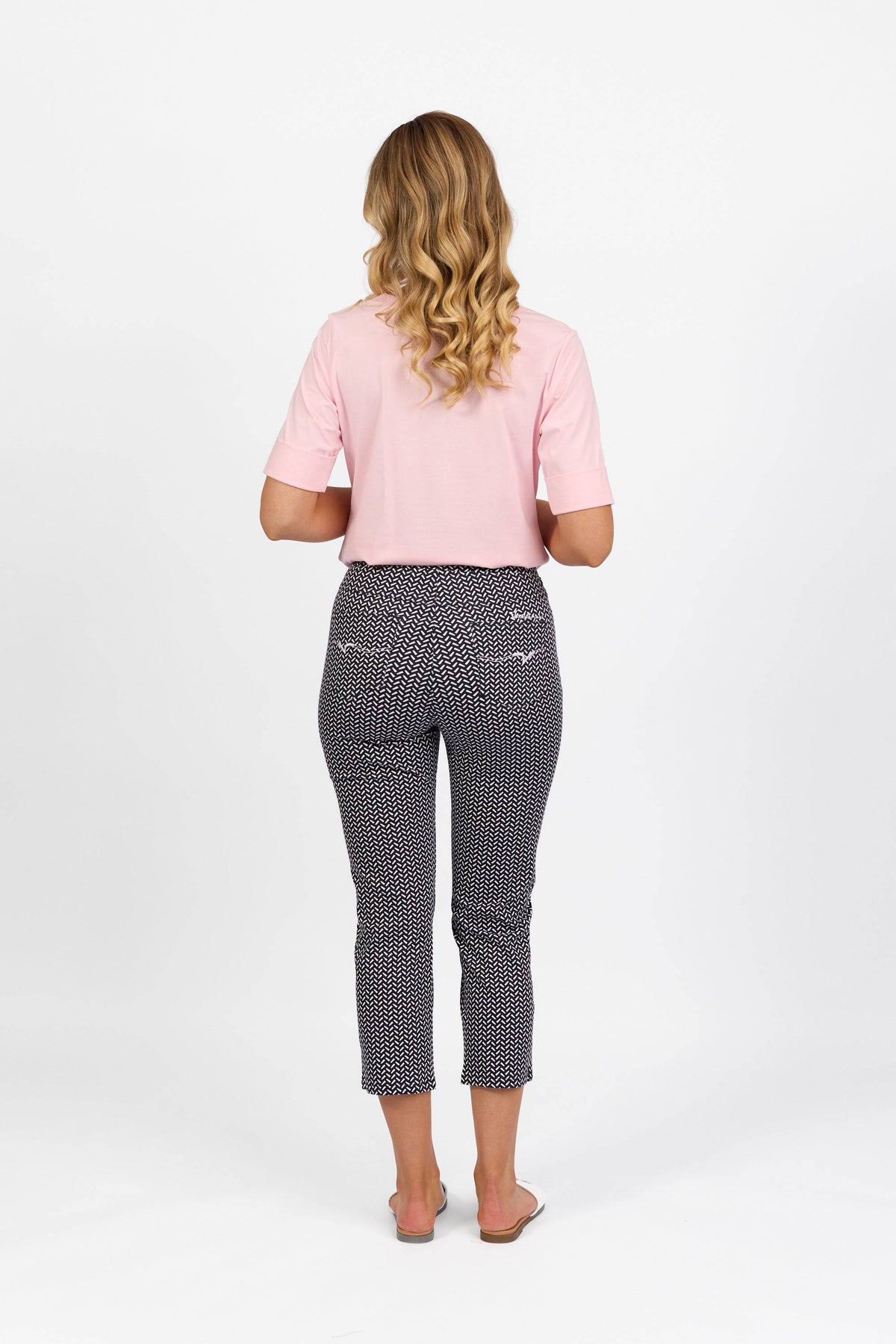 A woman with long, wavy blonde hair stands facing away. She wears a pink short-sleeve top and the Vassalli Sierra 7/8 Pant in Nori, crafted from stretch fabric, paired with flat sandals, against a plain white background.