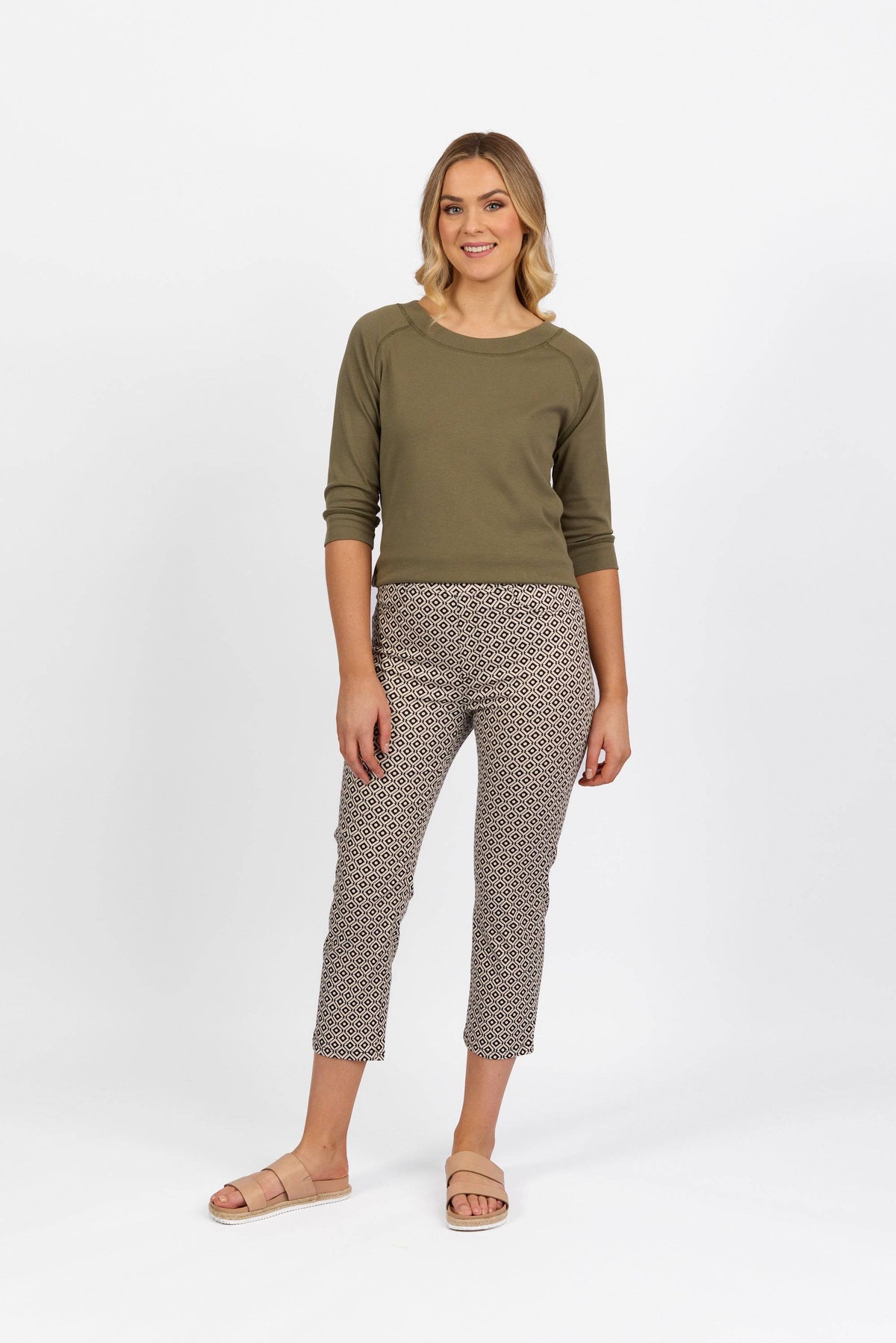 A woman smiles against a white background, wearing an olive green long-sleeve top, Vassalli Sierra 7/8 Pant in Sahara print, and light pink sandals.