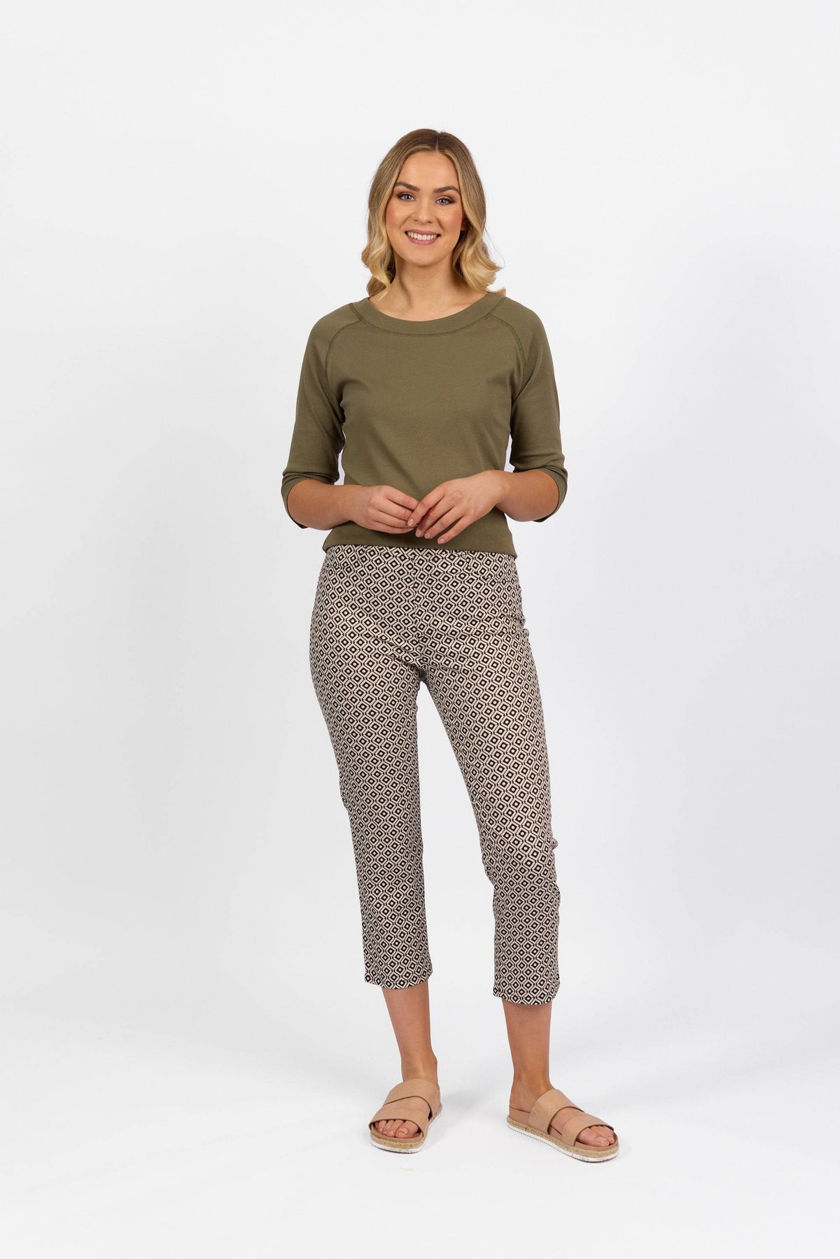 A woman stands smiling in a green three-quarter sleeve top, Vassalli Sierra 7/8 Pant - Sahara, and beige sandals, against a plain white background.