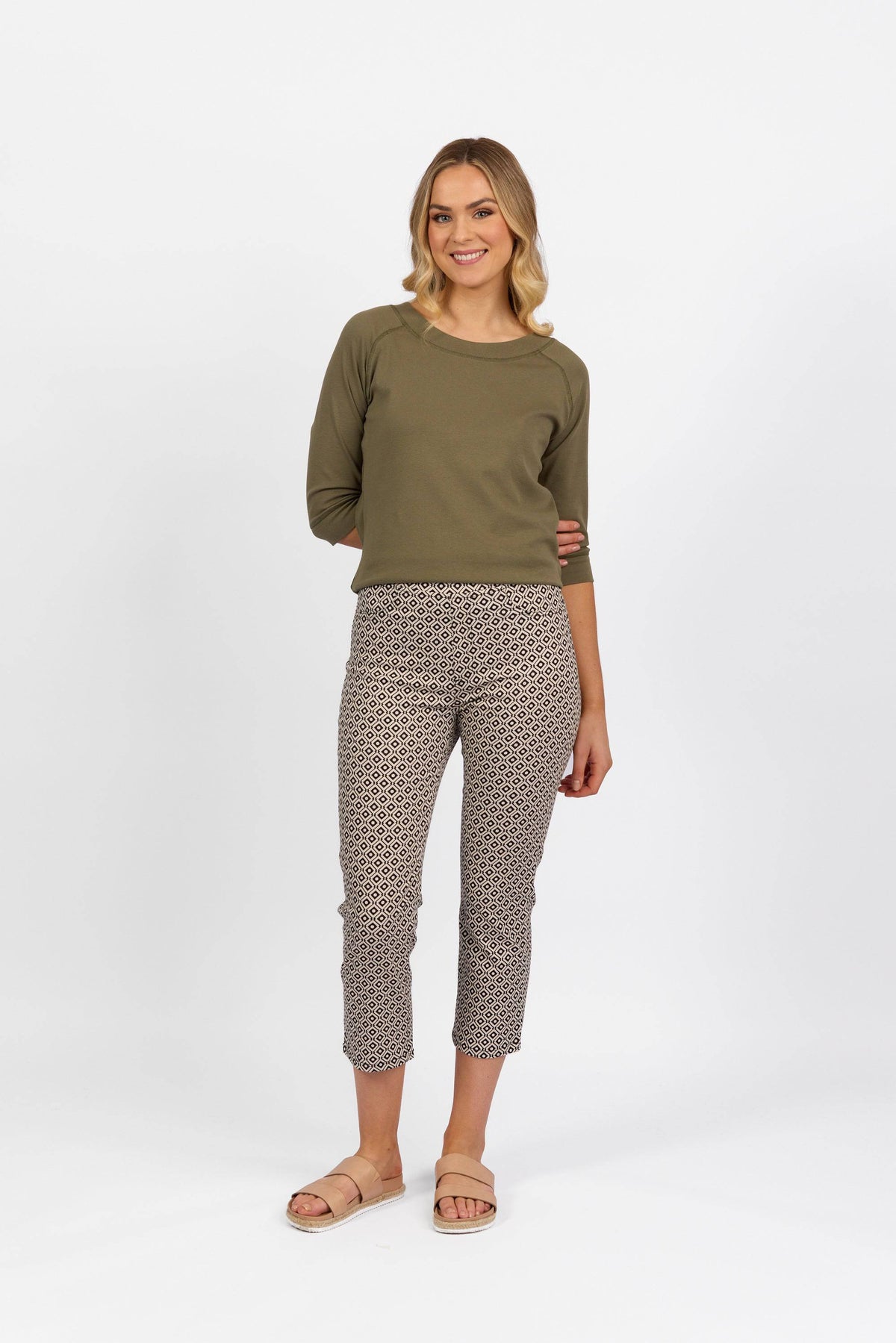A woman stands against a white background, smiling in an olive green long-sleeve top, beige sandals, and Vassalli Sierra 7/8 Pant - Sahara with an exclusive print. Her hair is styled in loose waves.