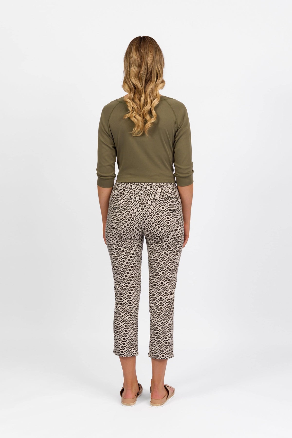 A person with long wavy blonde hair, facing away from the camera, wears an olive green top and Vassalli Sierra 7/8 Pant in Sahara print, paired with sandals against a plain white background.