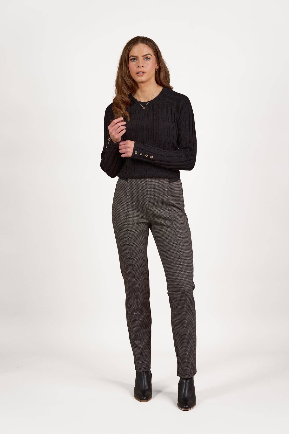A woman with long brown hair stands against a white background, wearing Vassalli's Everest Pull On - Jed gray checkered pants, a black ribbed long-sleeve sweater, and black ankle boots, with one hand raised near her chest.
