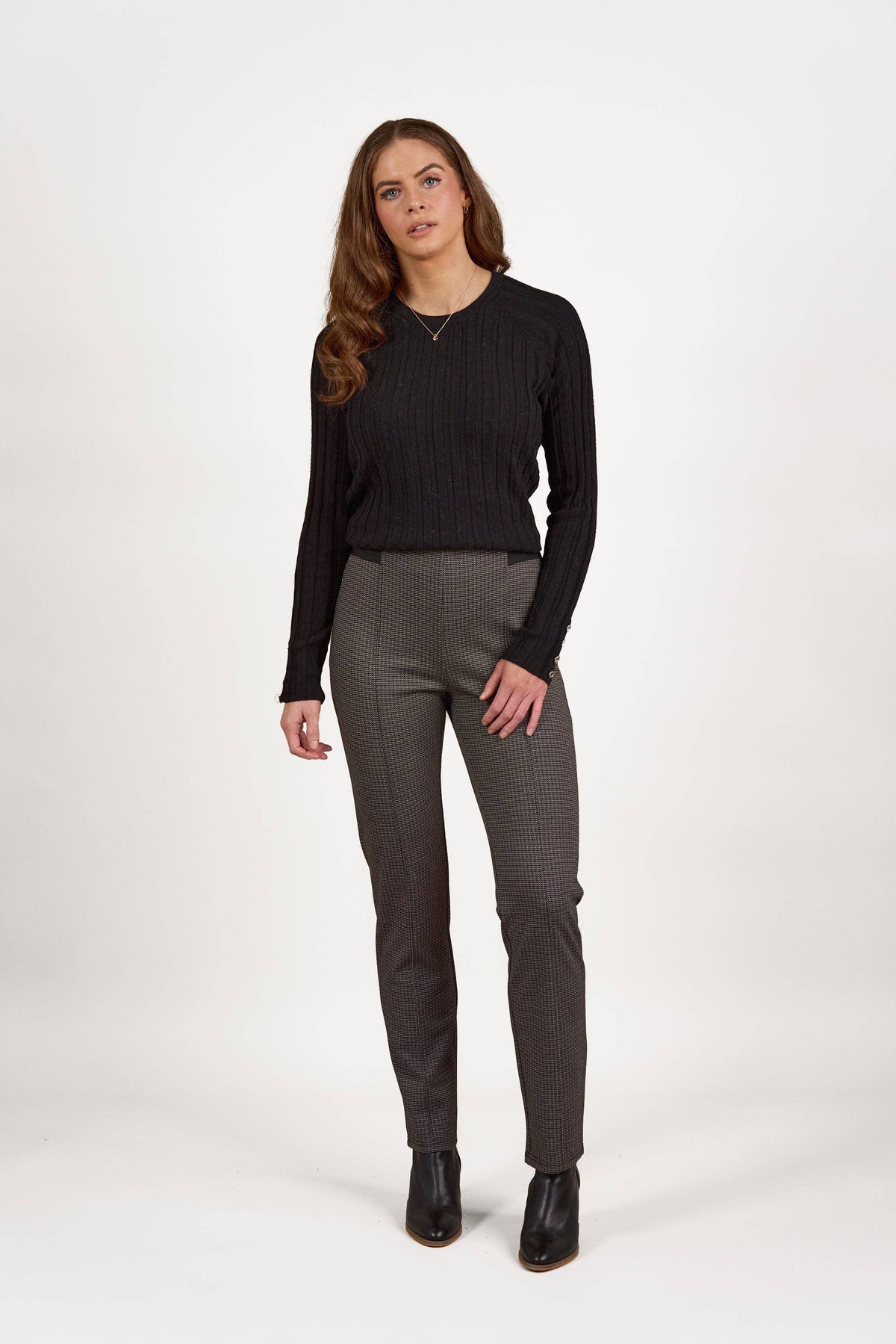 A woman with long brown hair wears the Vassalli Everest Pull On - Jed slim leg knit pants, a black ribbed sweater, and black ankle boots, standing against a plain white background.