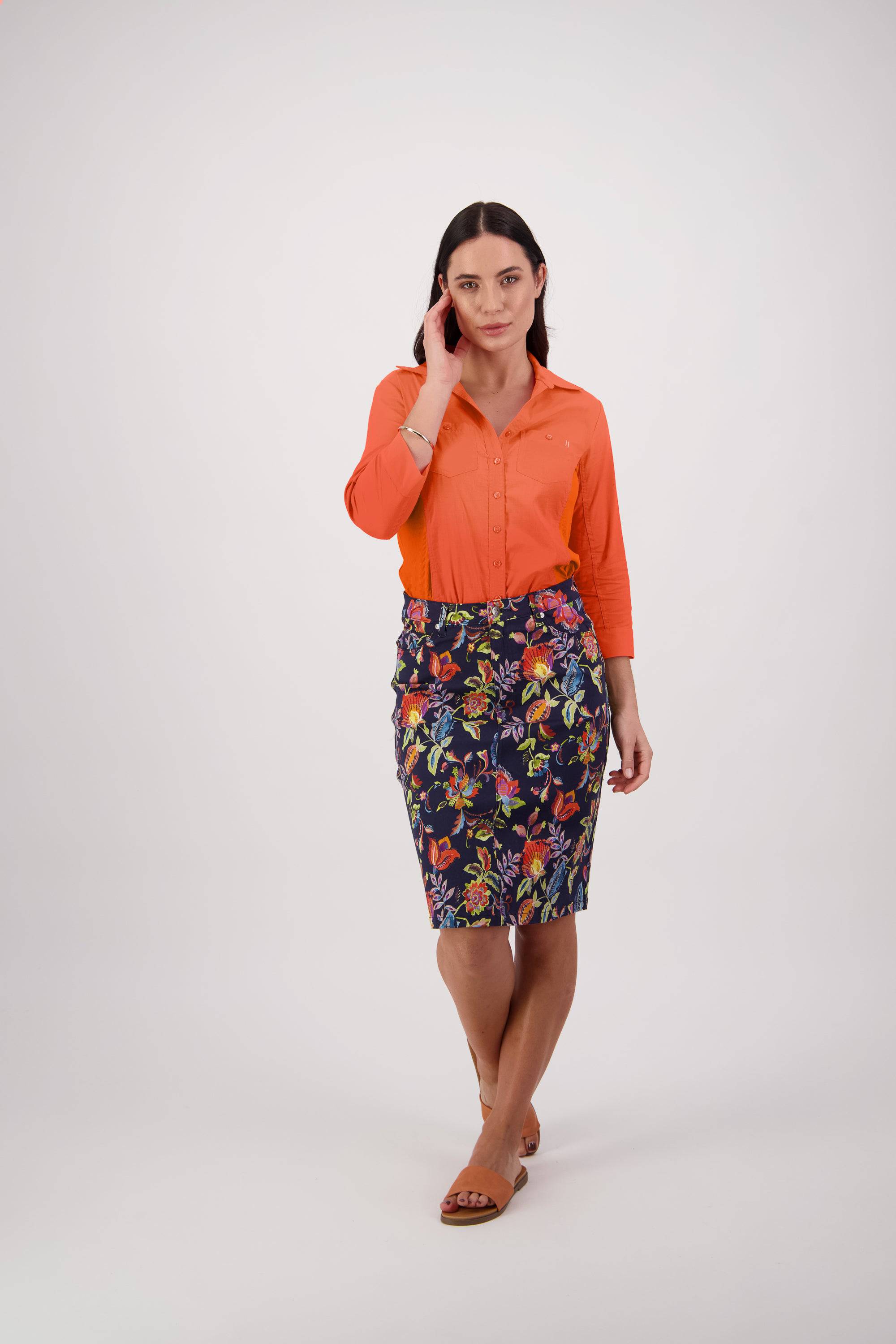 A woman with straight dark hair stands against a plain background wearing an orange button-up shirt, the Vassalli Poppy Skirt - Brazil, and brown flat sandals. She touches her face with one hand.