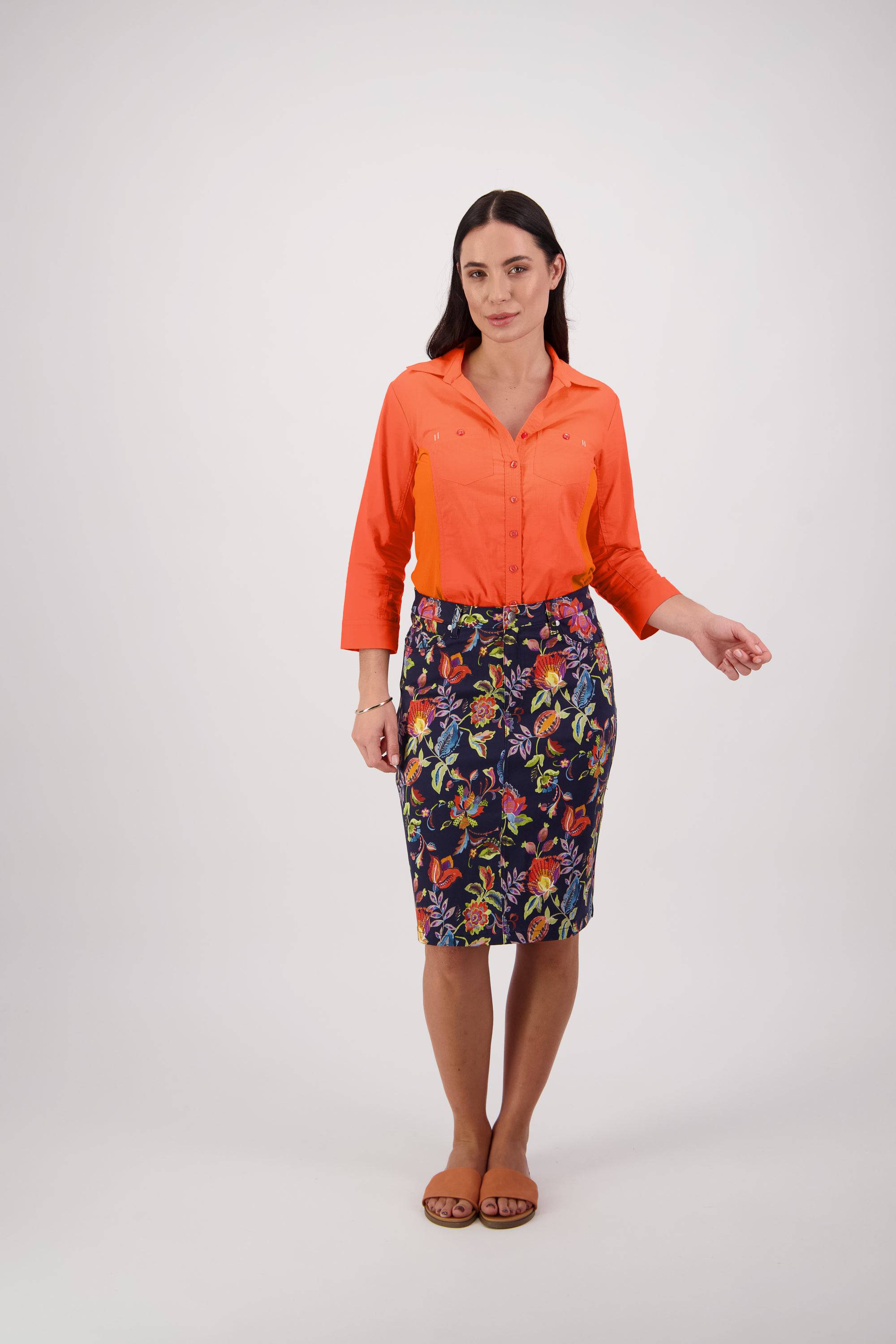 A woman stands against a plain white background wearing an orange button-up shirt, the Vassalli Poppy Skirt – Brazil, and flat brown sandals. She has long dark hair and three-quarter sleeves on her shirt.