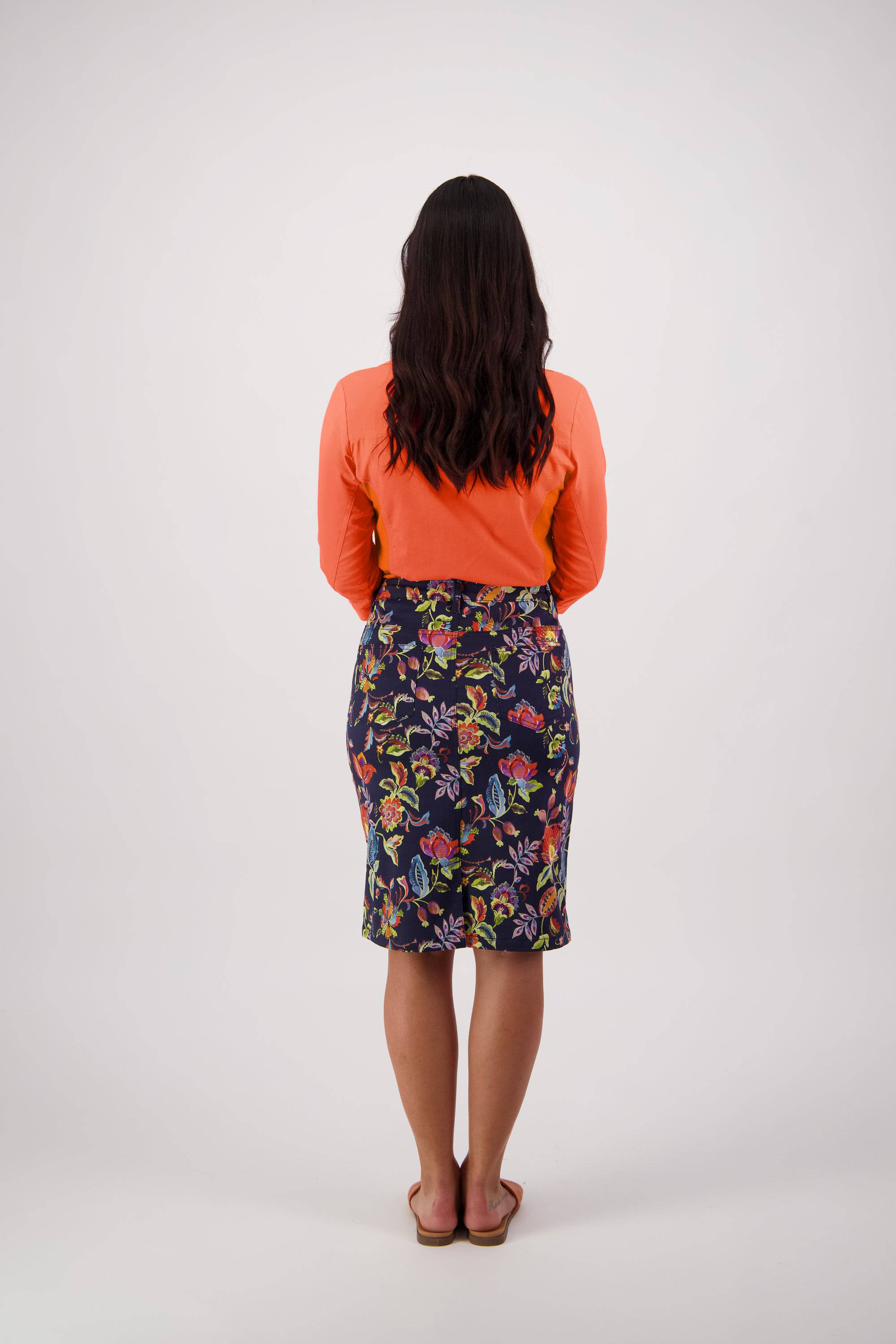 A woman with wavy dark hair, in an orange long-sleeve top and a Vassalli Poppy Skirt - Brazil with a centre back vent, stands barefoot facing away from the camera against a plain light background.