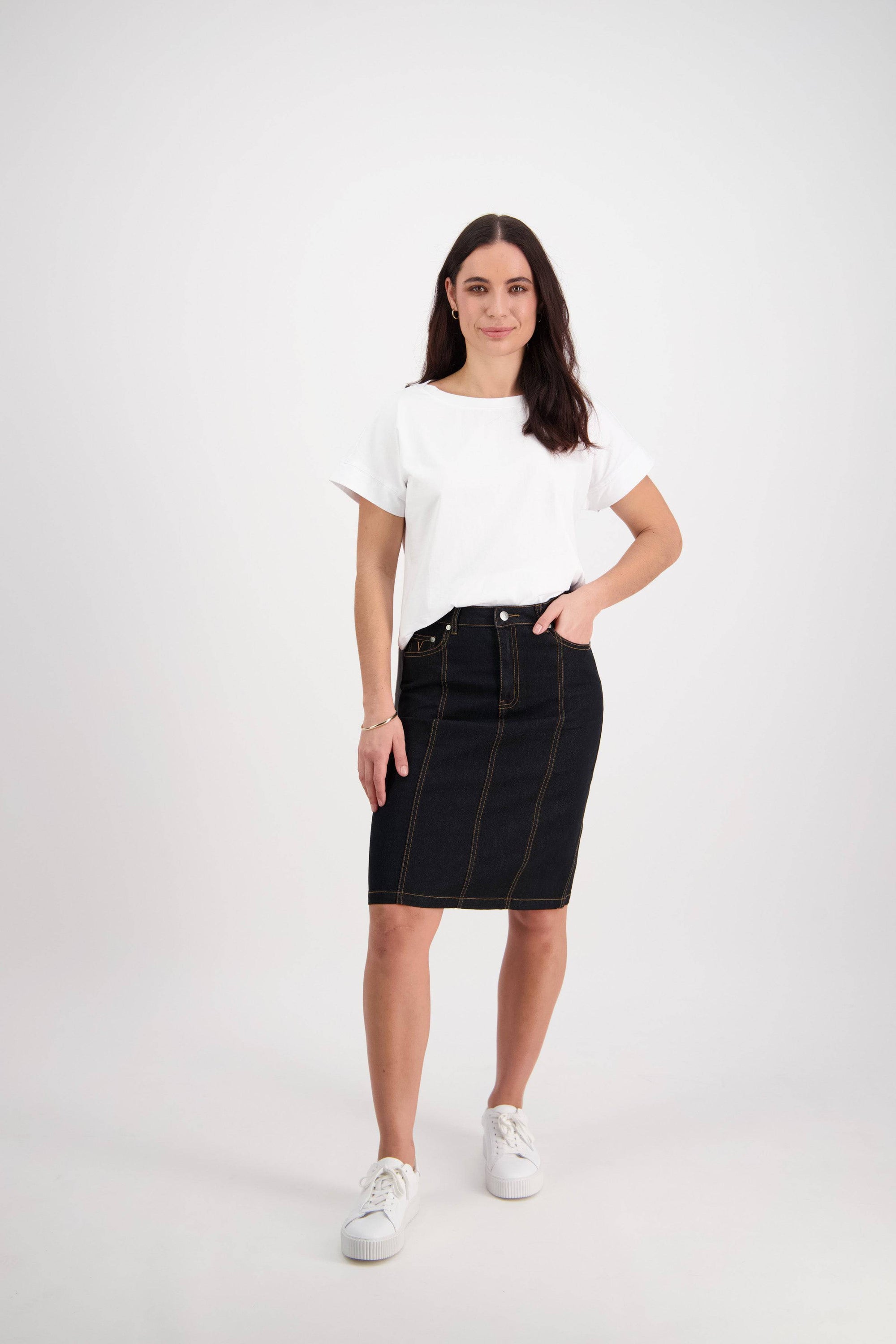 A woman with long dark hair stands against a white background, wearing a white t-shirt, the Vassalli Poppy Skirt in Black Denim, and white sneakers. One hand rests on her hip as she looks at the camera.