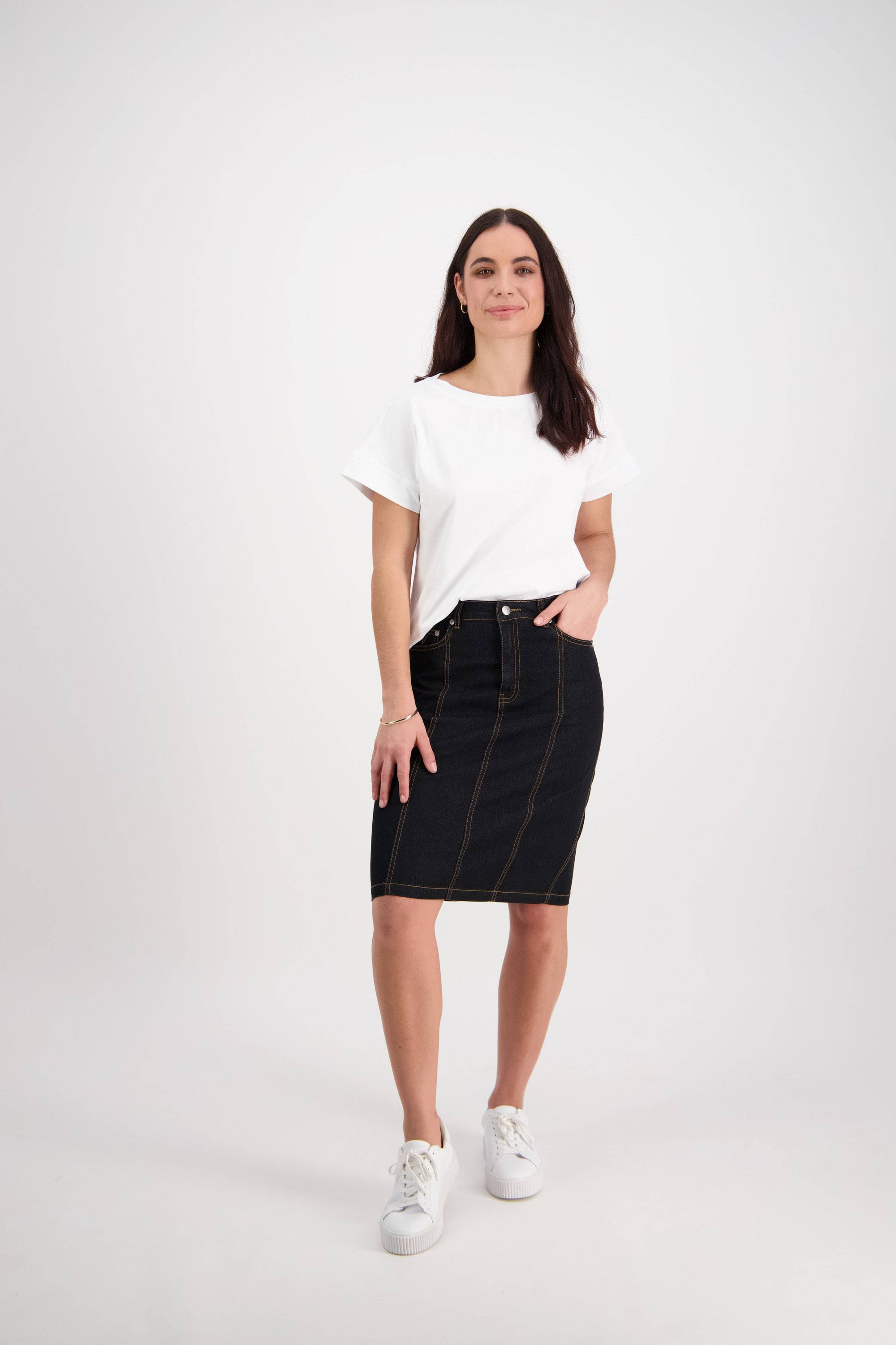 A woman stands against a plain white background, wearing a white t-shirt, the Vassalli Poppy Skirt in black denim, and white sneakers. She has long brown hair, one hand in her pocket, and is smiling softly.