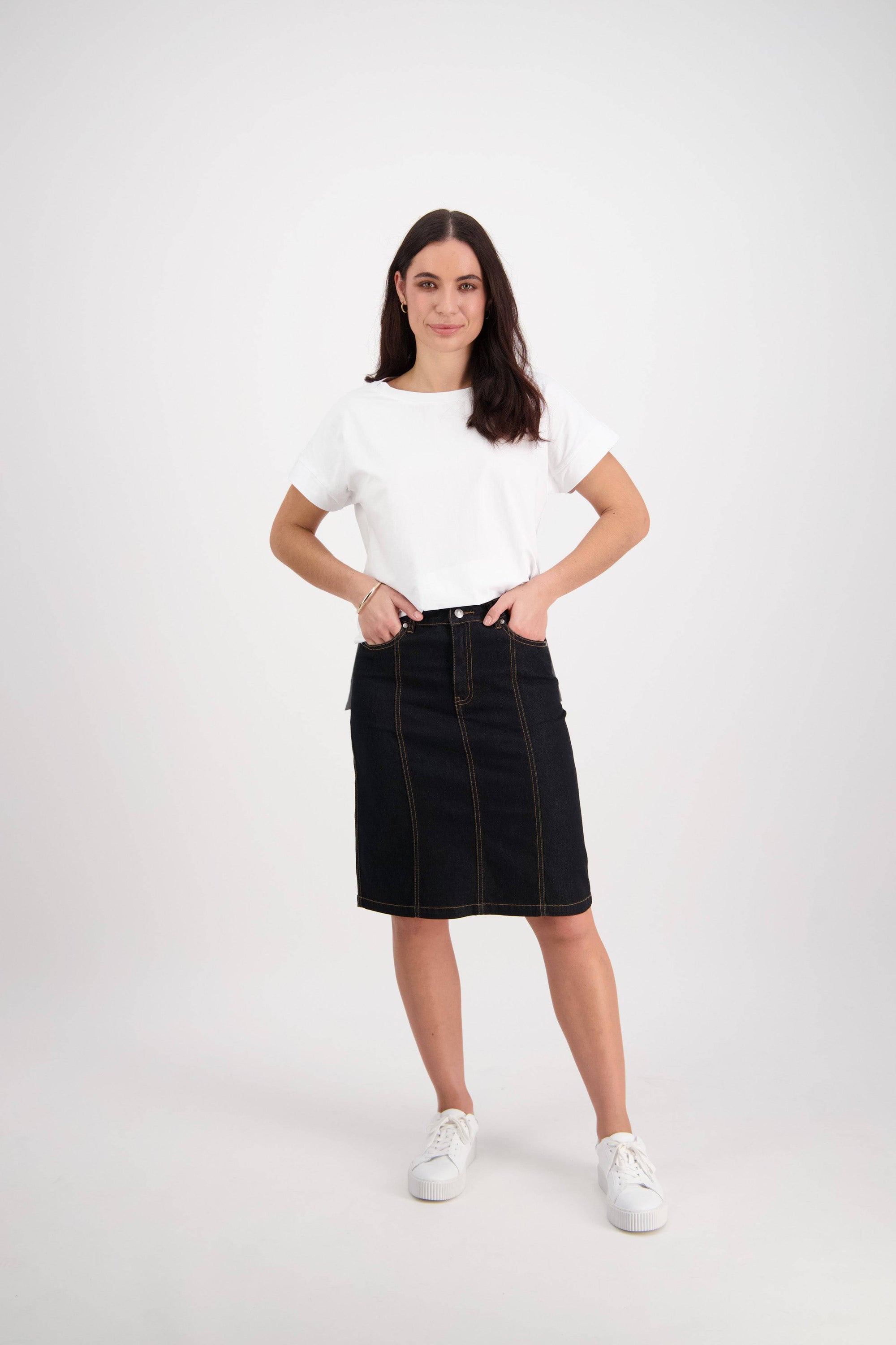 A woman stands with hands on hips, wearing a white t-shirt, the Vassalli Poppy Skirt in black denim featuring contrast stitch details, and white sneakers, set against a plain white background.