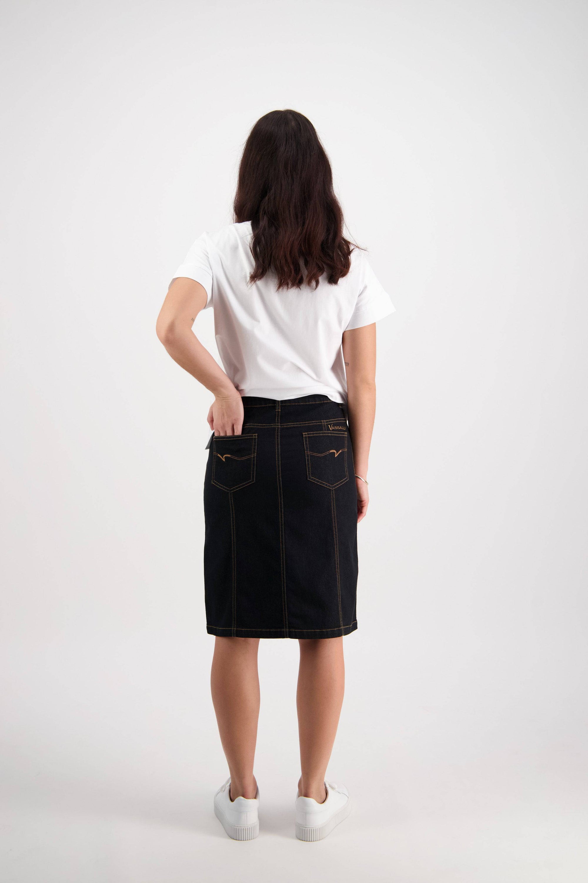 A woman stands facing away against a plain white background, wearing a white t-shirt, white sneakers, and the Vassalli Poppy Skirt in black denim with contrast stitching, with one hand in her back pocket.