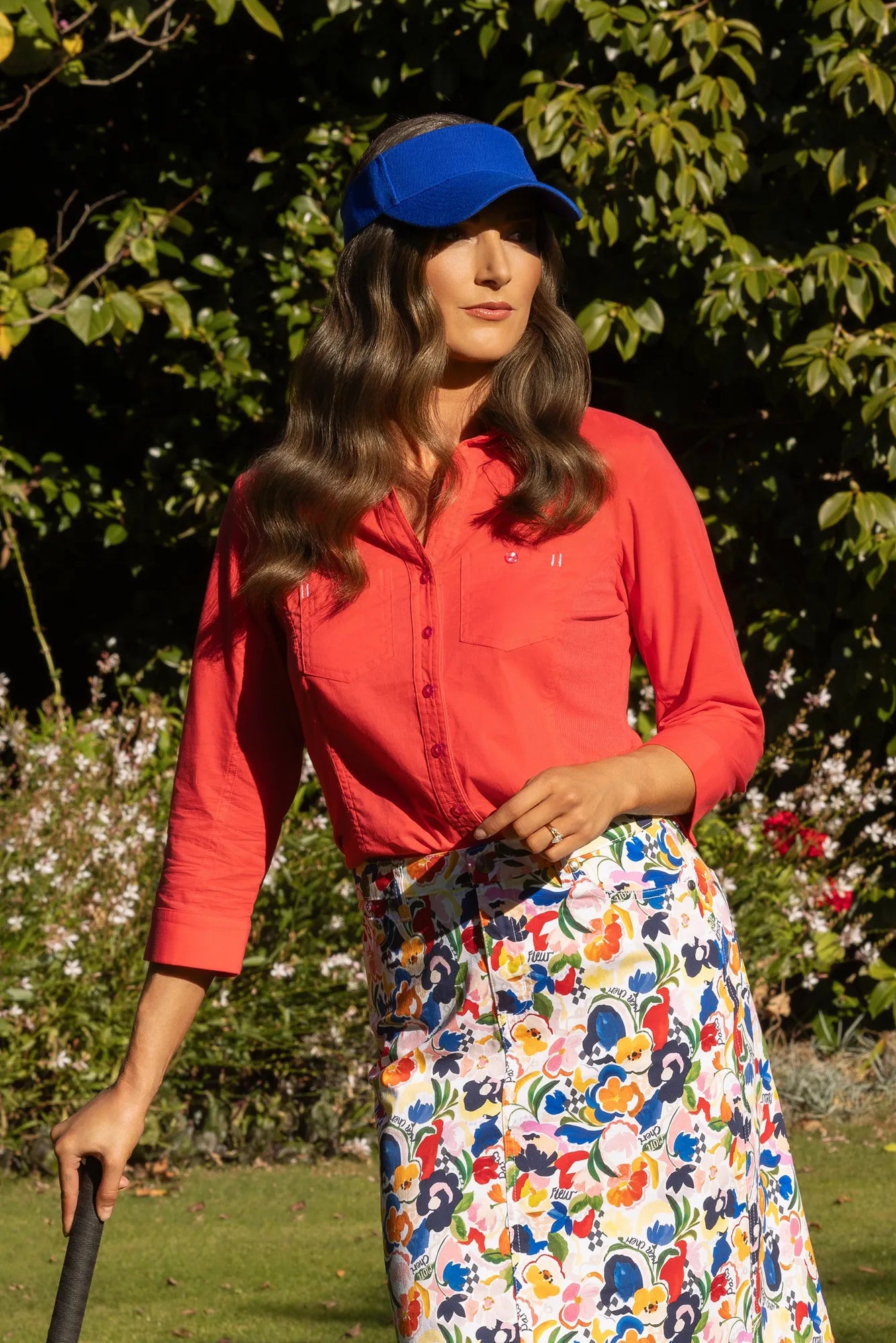 A person wearing a blue cap, the Vassalli Isla Shirt in Raspberry, and a colorful floral skirt stands outdoors in a garden, holding a cane.