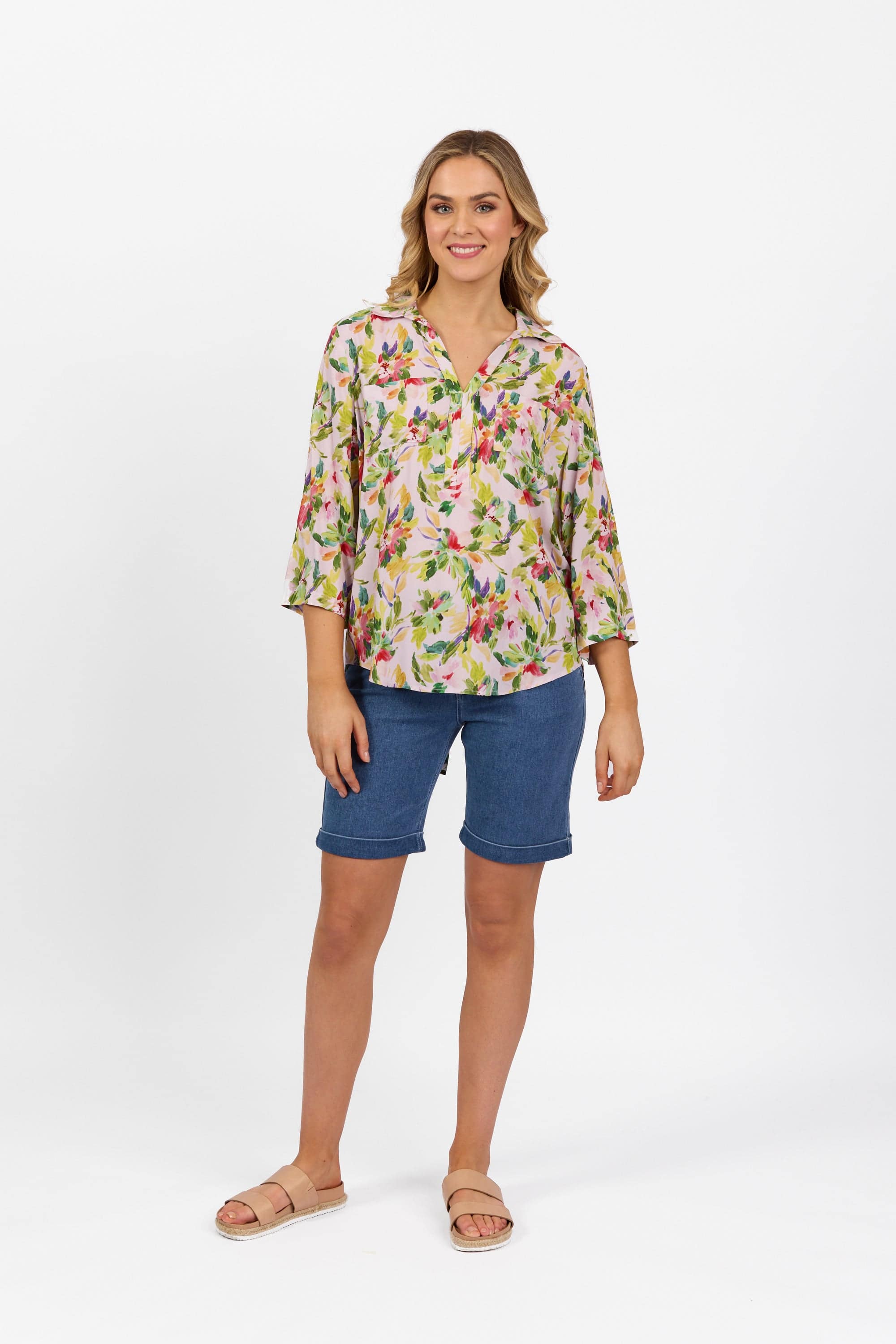 A woman stands smiling against a plain white background, wearing the Vassalli Placket Shirt - Capri, denim knee-length shorts, and sandals.