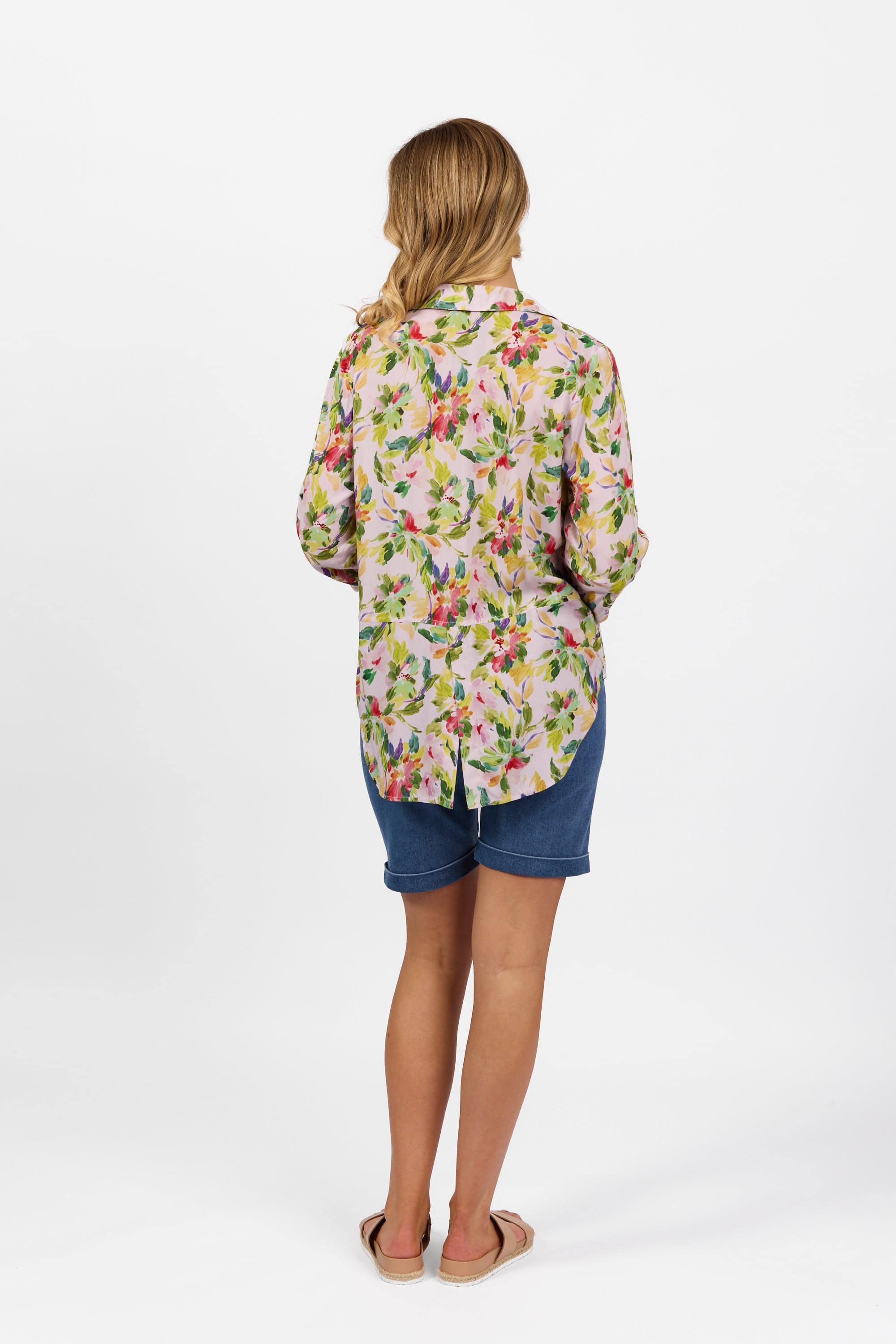 A woman stands with her back to the camera, wearing the Vassalli Placket Shirt - Capri, blue knee-length shorts, and beige sandals against a plain white background.