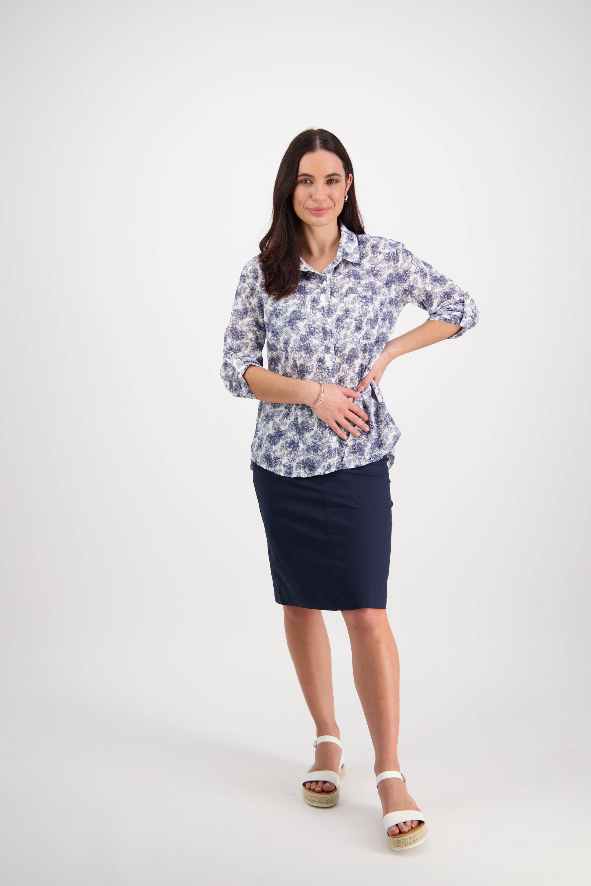 A woman with long dark hair smiles against a white background, wearing the Vassalli Crinkle Shirt - Denim Floral, a navy skirt, and white sandals. One hand rests on her hip and the other on her stomach.