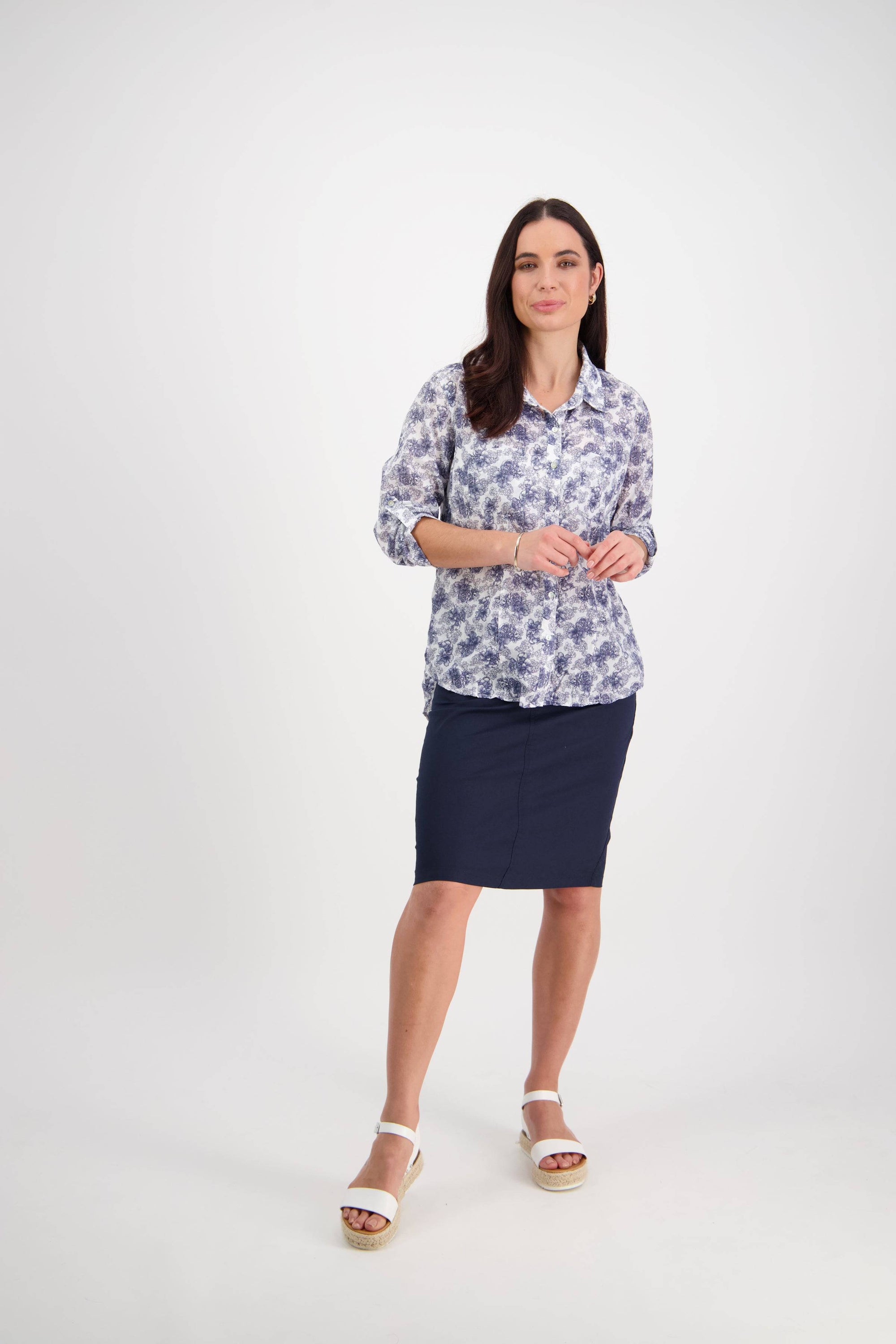 A woman with long dark hair and a slight smile stands against a plain white background, wearing the Vassalli Crinkle Shirt - Denim Floral, a navy knee-length skirt, and white platform sandals.