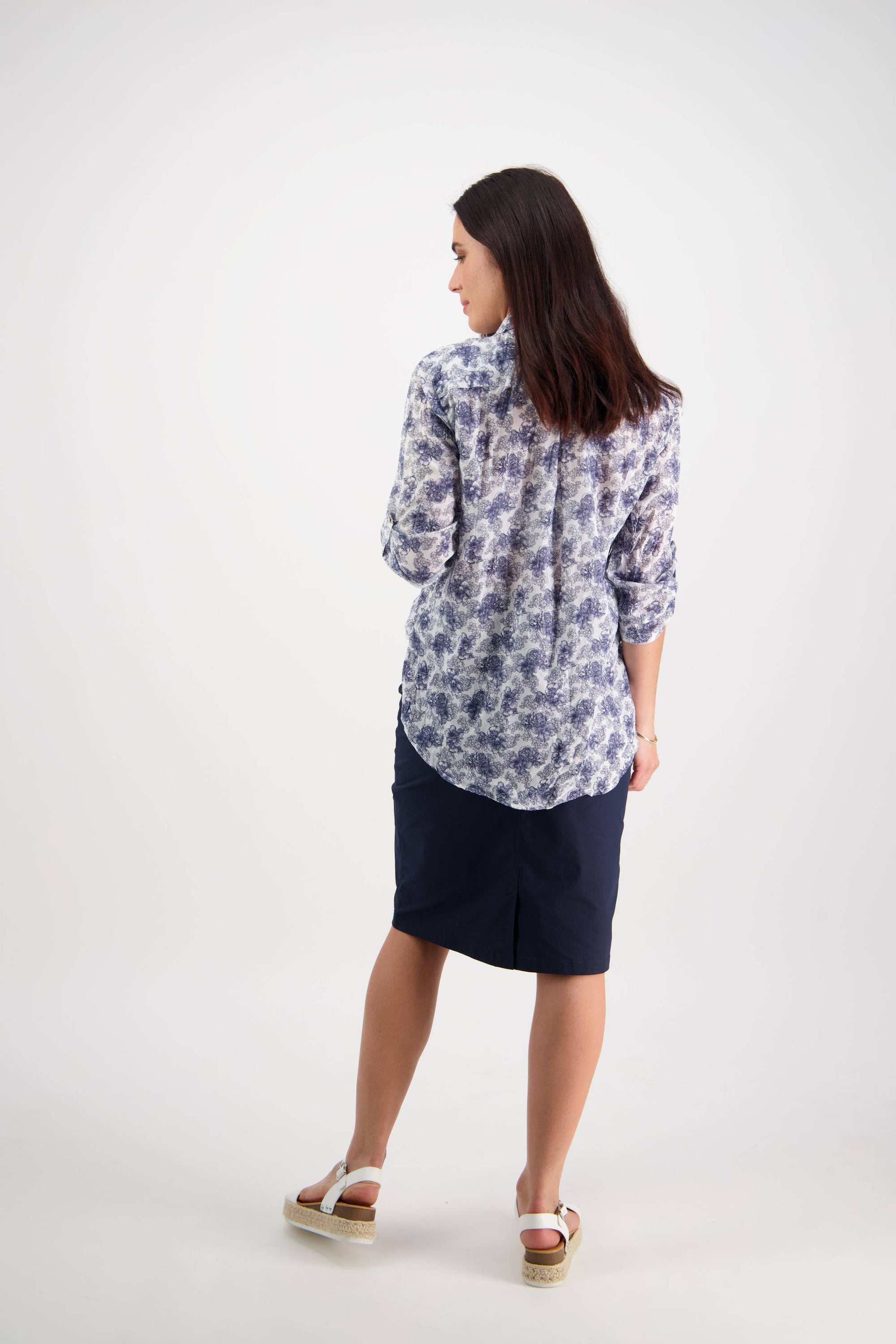 A woman with dark hair faces away, wearing the Vassalli Crinkle Shirt - Denim Floral with long sleeves, paired with a navy knee-length skirt and white sandals, standing in a light, plain studio.