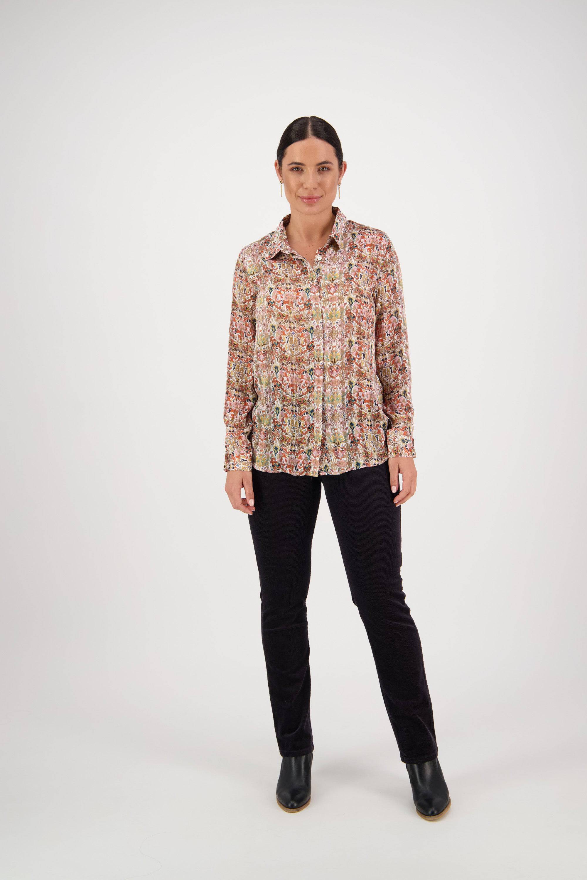 A woman faces forward in the Vassalli Ballot Shirt - Eden, which features colorful floral patterns and subtle gold lurex stripes, styled with black pants and ankle boots against a plain white background.