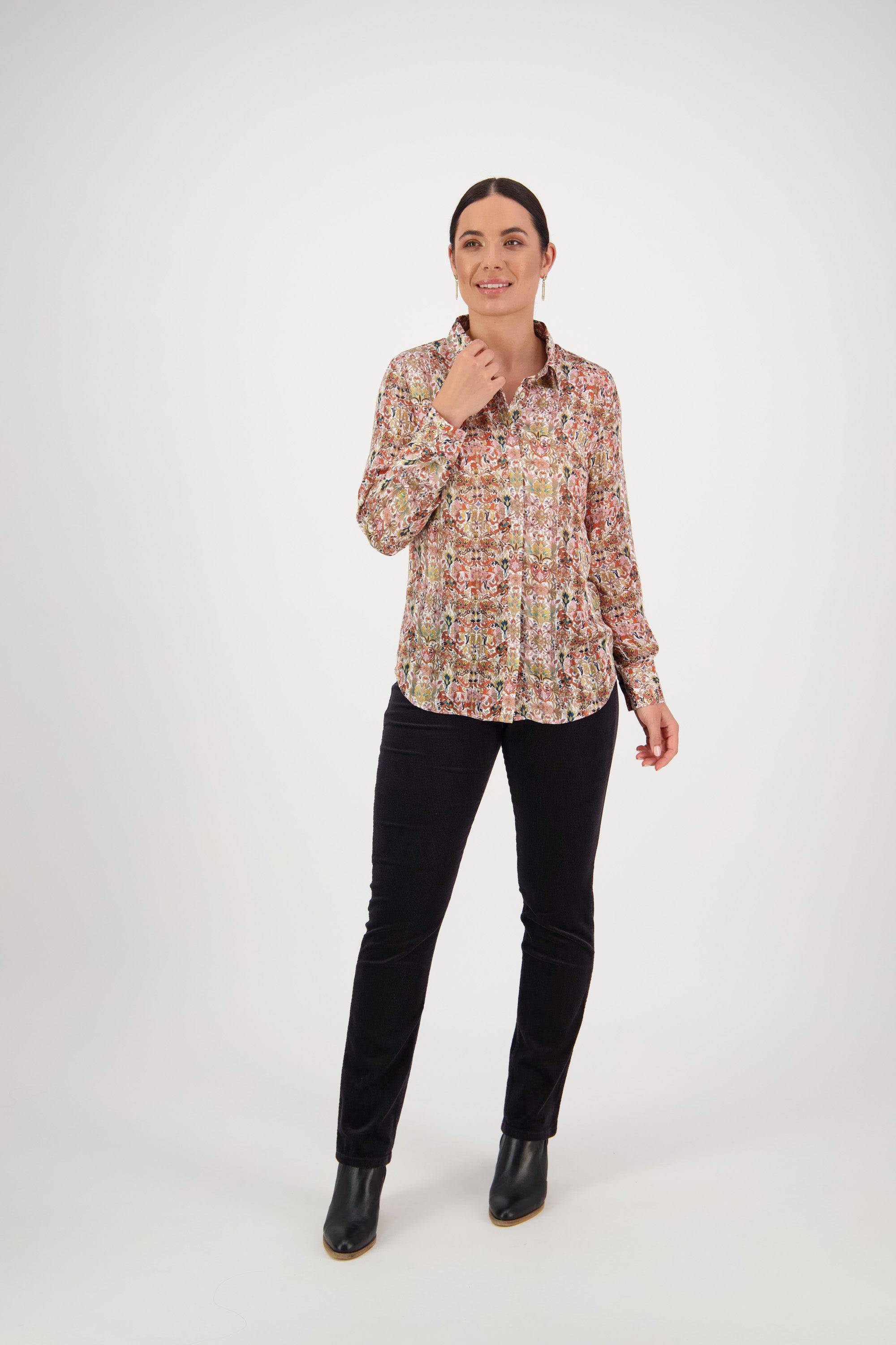 A woman stands against a plain white background, smiling as she wears the Vassalli Ballot Shirt - Eden with black pants and ankle boots. One hand touches her collar while the other rests by her side.