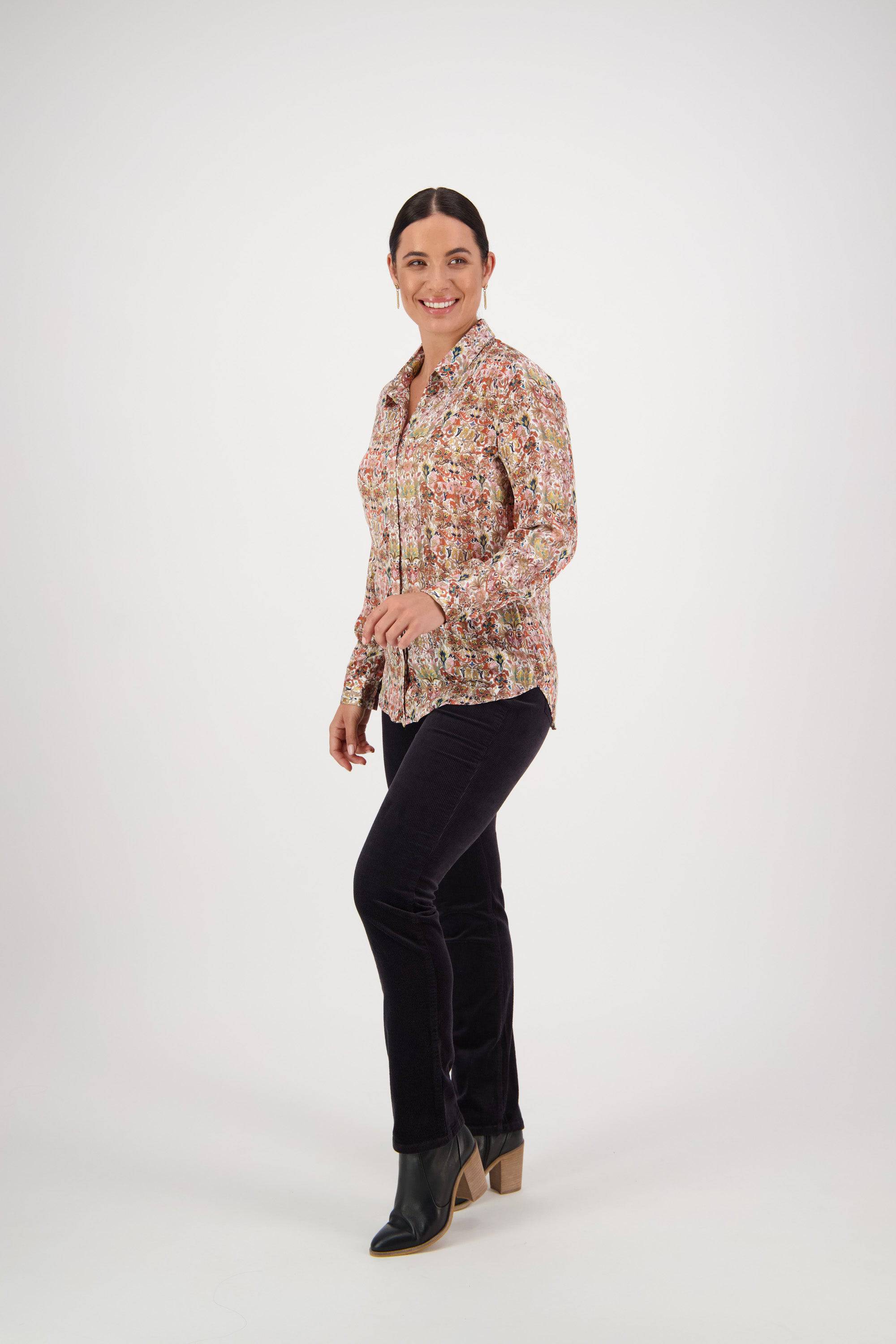 A woman with dark hair is smiling and walking, wearing the Vassalli Ballot Shirt - Eden, black pants, and heeled ankle boots, standing against a plain white background.