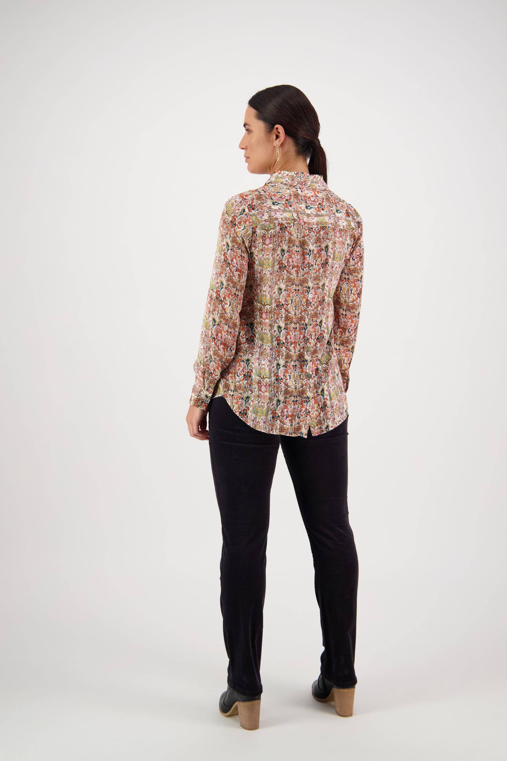 A woman with her dark hair in a low ponytail faces left, wearing the Vassalli Ballot Shirt - Eden, featuring floral patterns and gold lurex stripes, paired with black pants and shoes against a white background.
