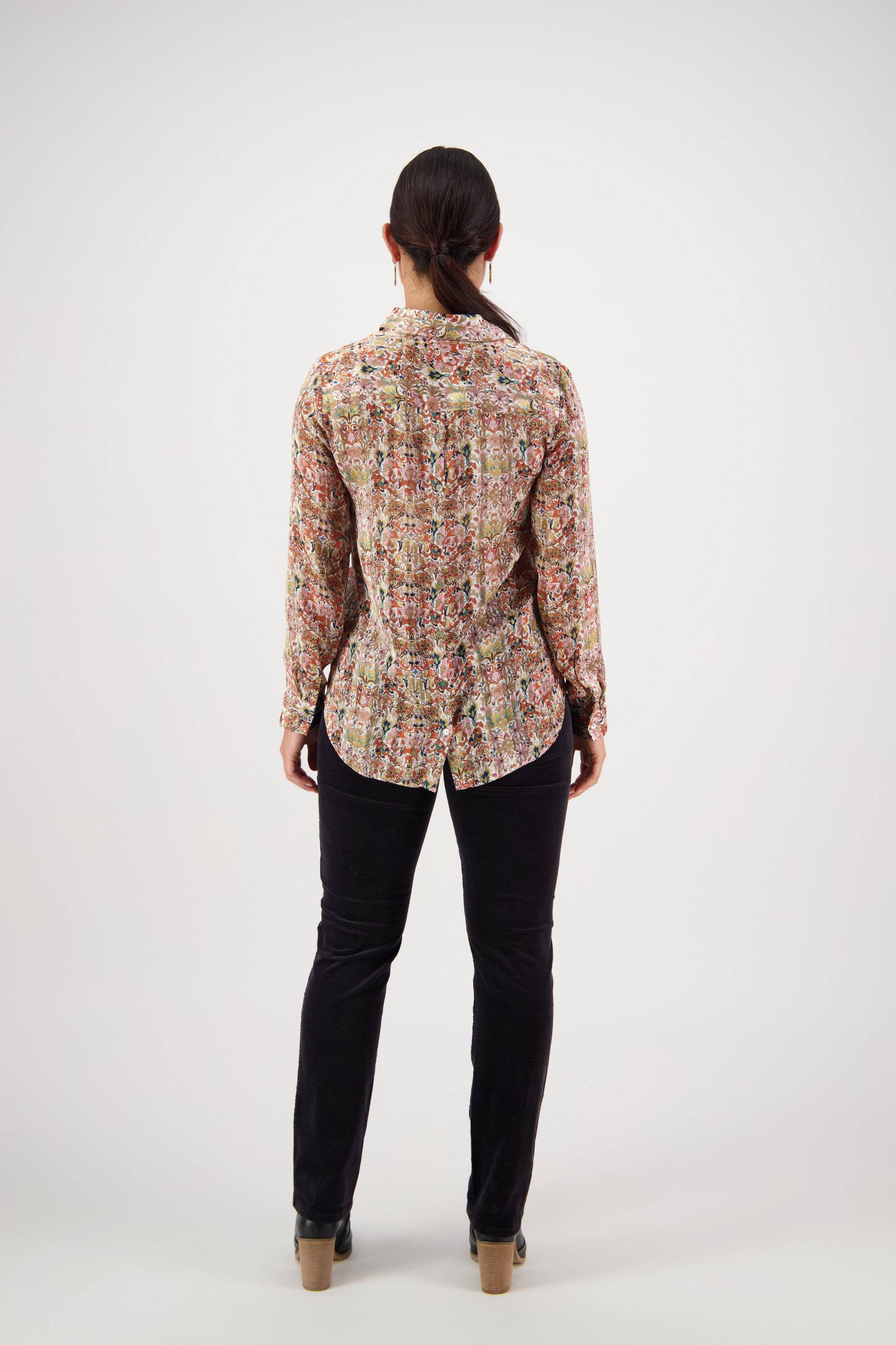 A person stands facing away from the camera in the Vassalli Ballot Shirt - Eden, featuring gold lurex stripes, paired with dark pants and heeled shoes, hair tied back in a ponytail against a plain light background.