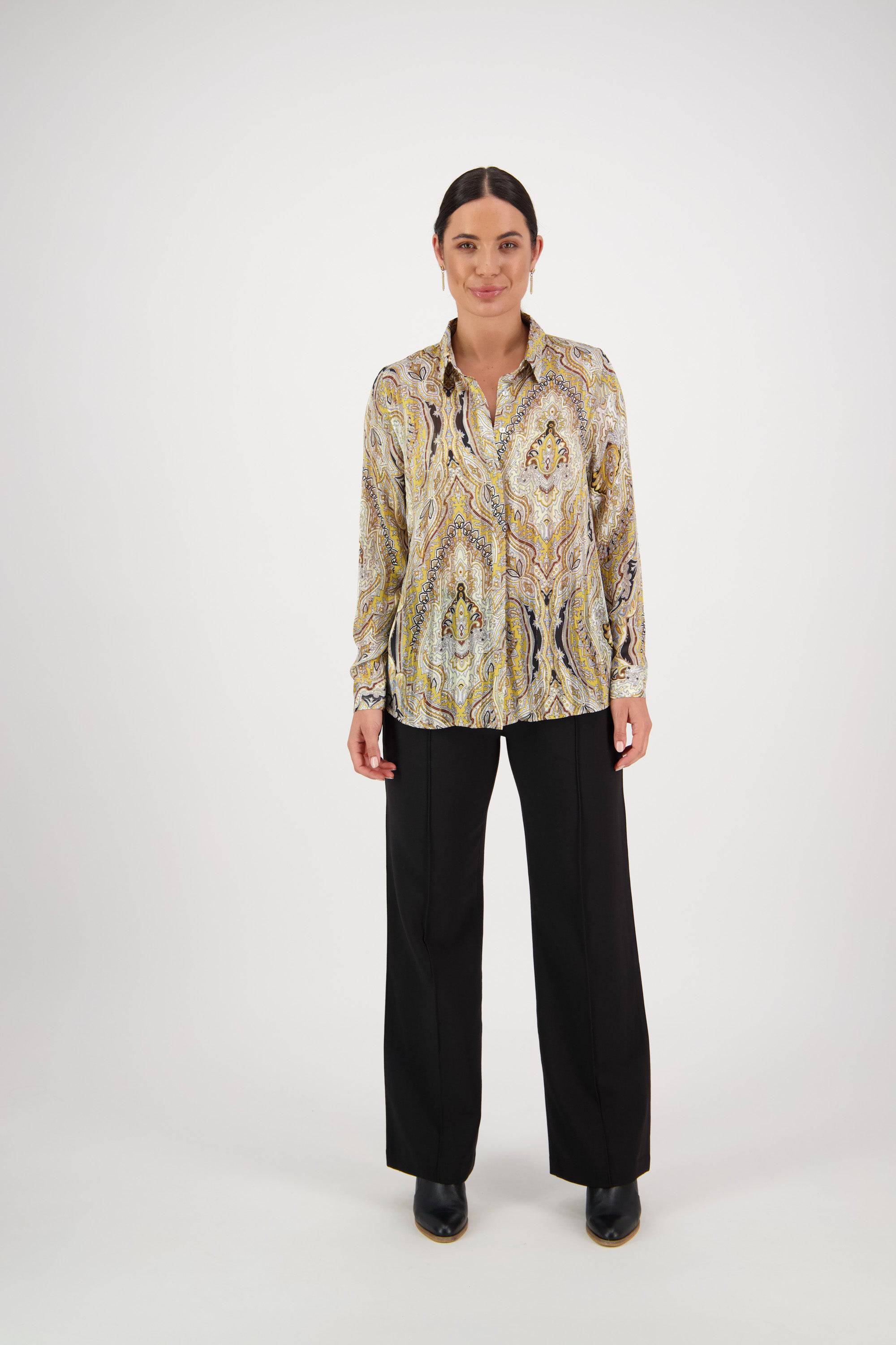 A woman with dark hair pulled back stands against a white background, wearing the Vassalli Ballot Shirt - Majestic with gold lurex stripes and semi-sheer fabric, paired with black wide-leg pants and black shoes. She smiles slightly.