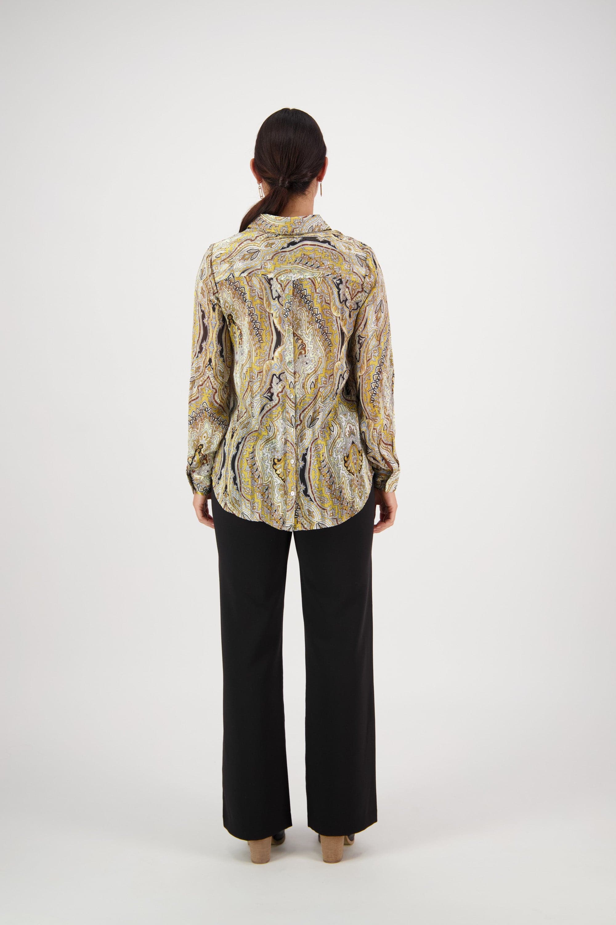 A person with long dark hair faces away from the camera, wearing the Vassalli Ballot Shirt - Majestic in patterned yellow, black, and white semi-sheer fabric with gold lurex stripes, paired with black pants against a plain white background.