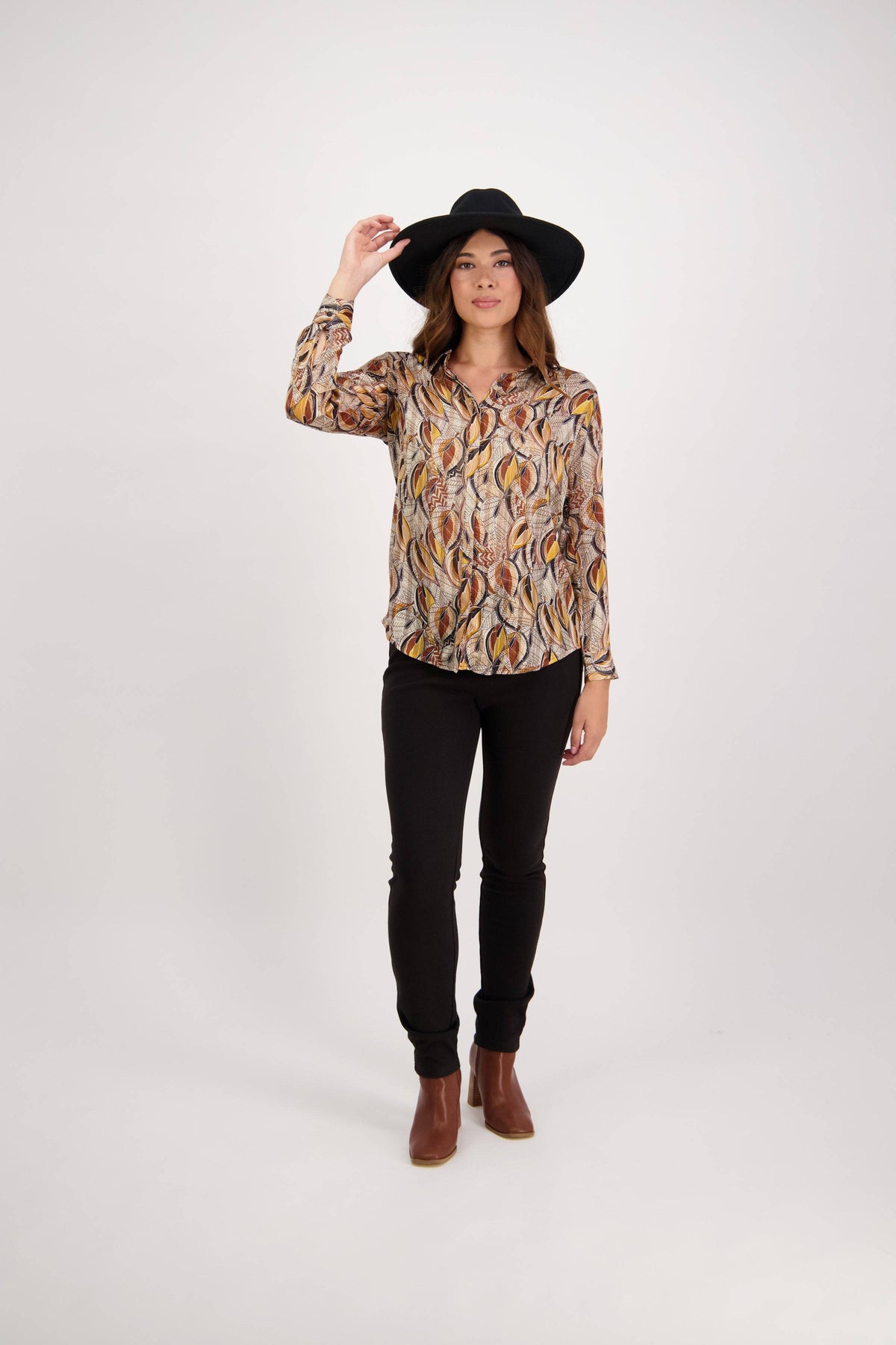 A woman stands against a plain white background, wearing the Vassalli Ballot Shirt - Willow with gold lurex stripes, black pants, brown ankle boots, and a black wide-brim hat. She touches the hat's brim and looks forward.