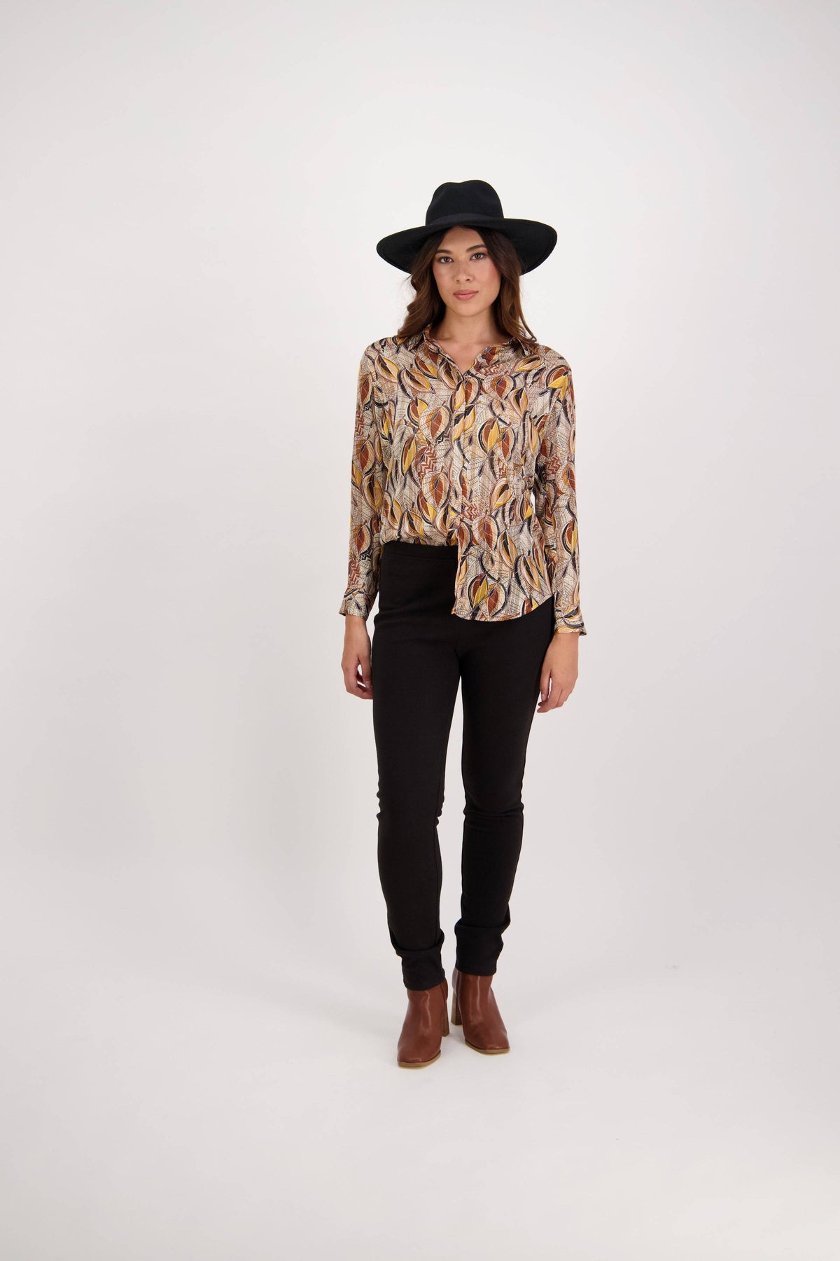 A woman wearing a wide-brimmed black hat, Vassalli's Ballot Shirt - Willow with gold lurex stripes, black pants, and brown ankle boots stands against a plain white background.