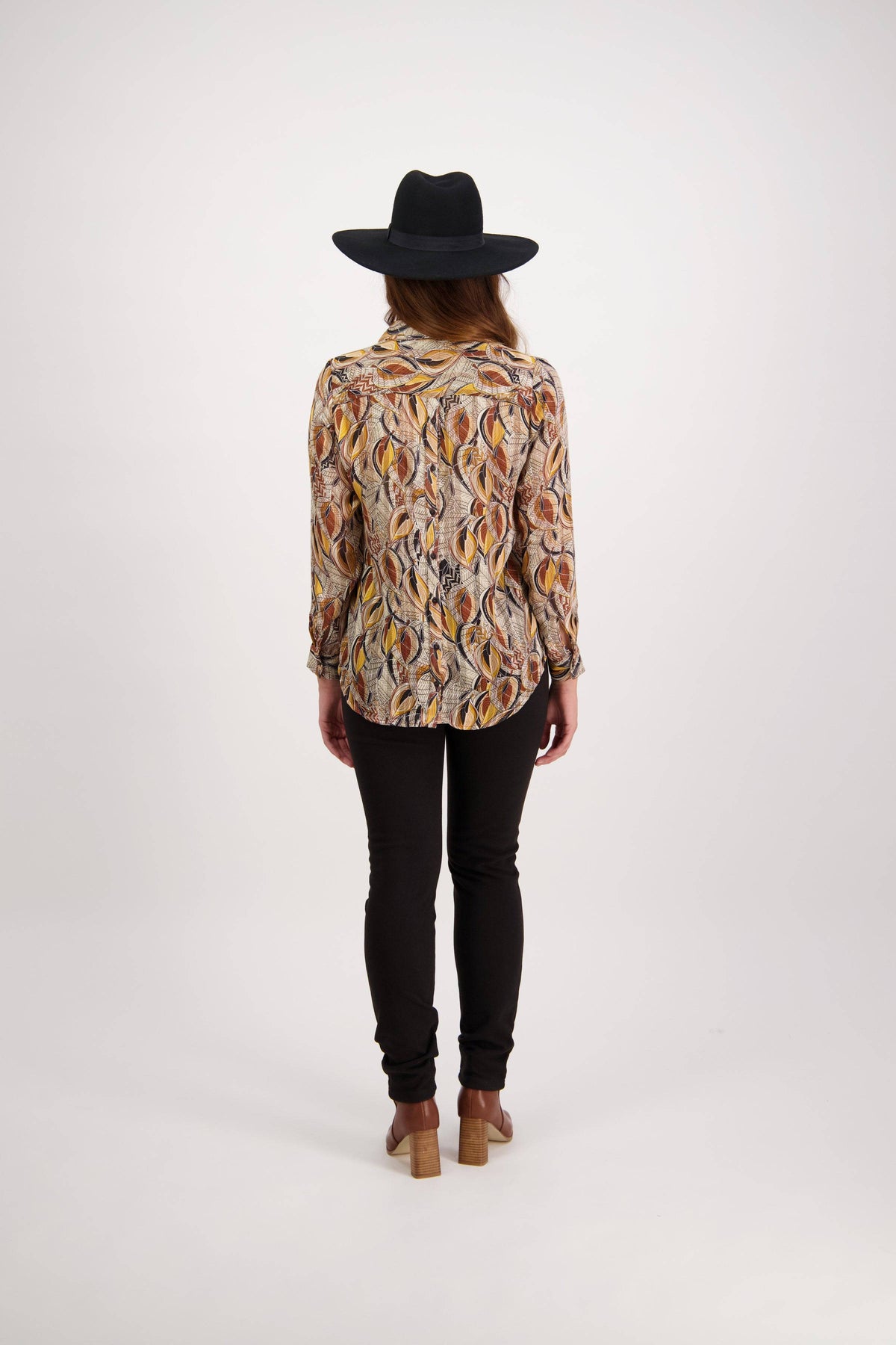 A person wears a black wide-brim hat, black pants, the Vassalli Ballot Shirt - Willow with gold lurex stripes in semi-sheer fabric, and tan heeled shoes, standing with their back to the camera against a plain white background.
