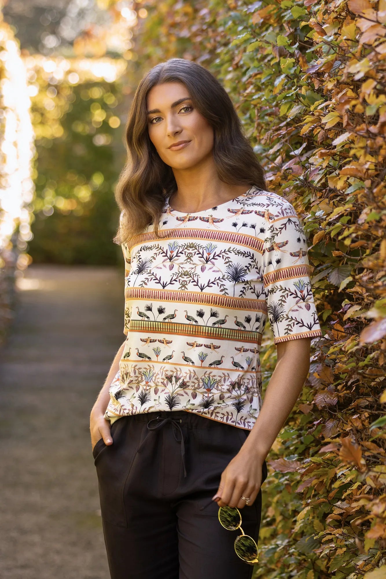 A woman stands outdoors beside a leafy hedge, wearing the Vassalli Carmen Top - Arli, made from soft rayon-elastane fabric and styled with dark pants. She holds sunglasses in one hand and gazes at the camera as gentle sunlight filters in behind her.
