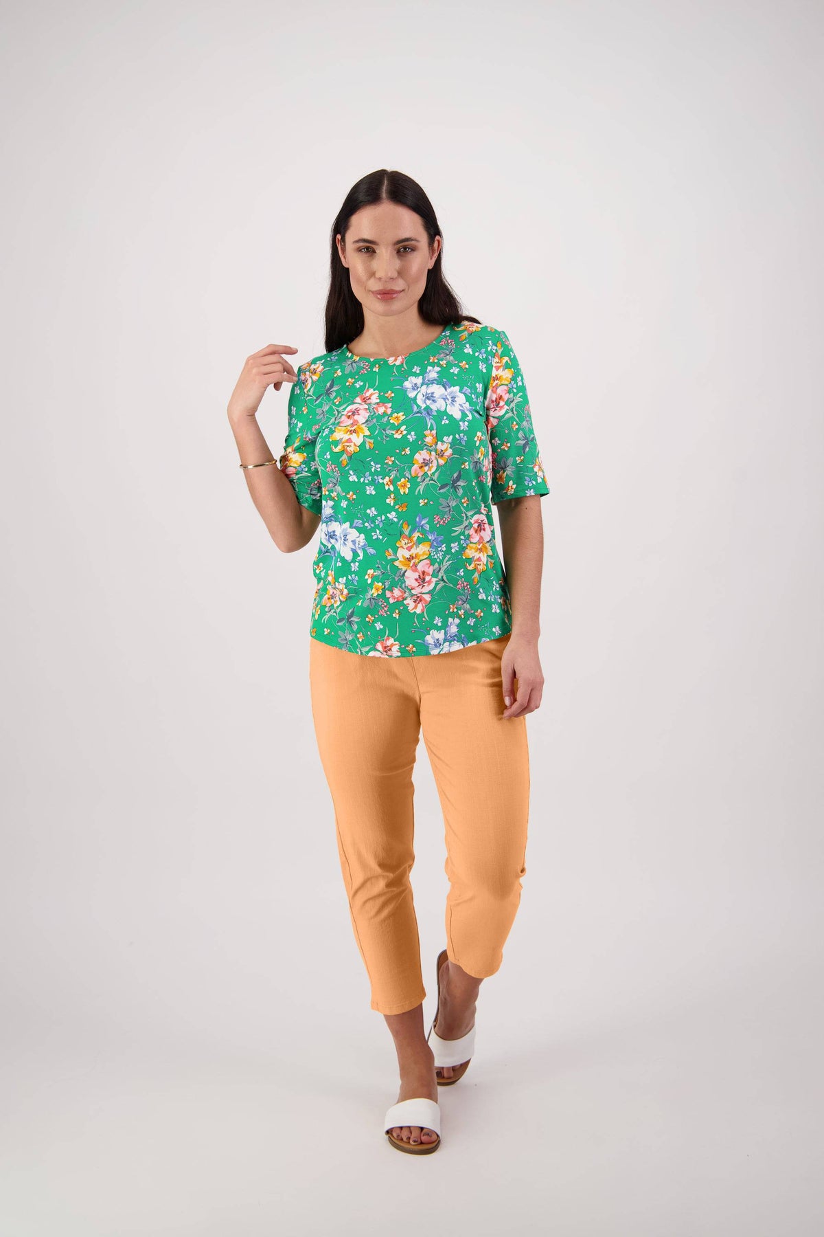 A woman with long brown hair and a neutral expression stands against a white background in the Vassalli Carmen Top - Lush, styled with yellow-orange pants and white open-toe sandals, one hand raised near her shoulder.