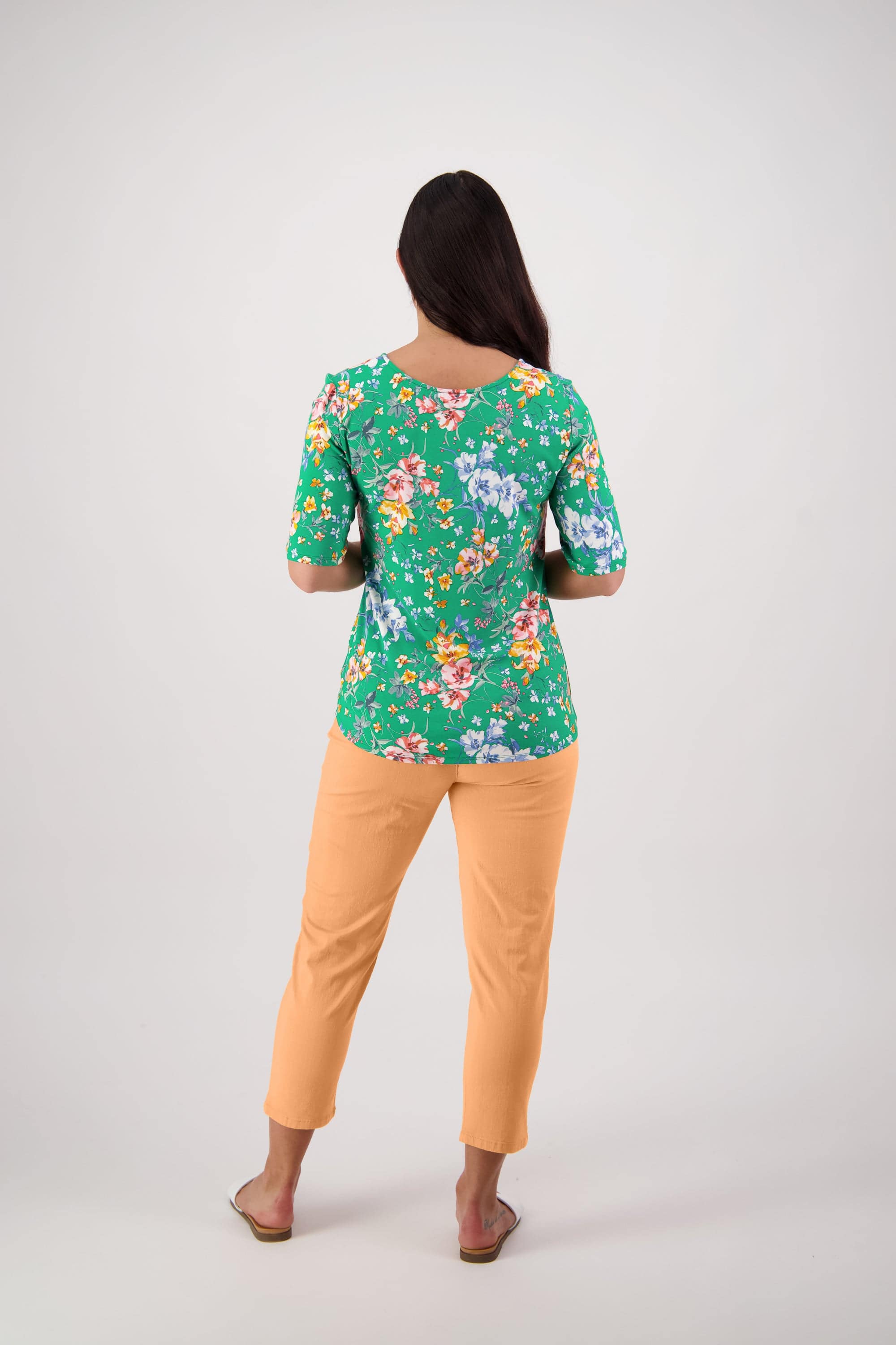 A woman with long dark hair faces away, wearing the Vassalli Carmen Top - Lush (a green floral 3/4 sleeve knit), peach pants, and sandals against a plain light background.