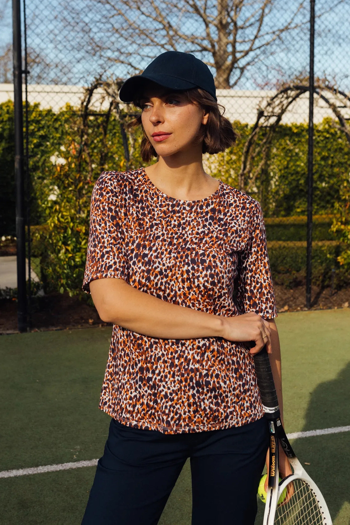 A woman wearing the Carmen Top by Zella from Vassalli and a black cap stands on a tennis court holding a tennis racket, with greenery and a chain-link fence in the background.