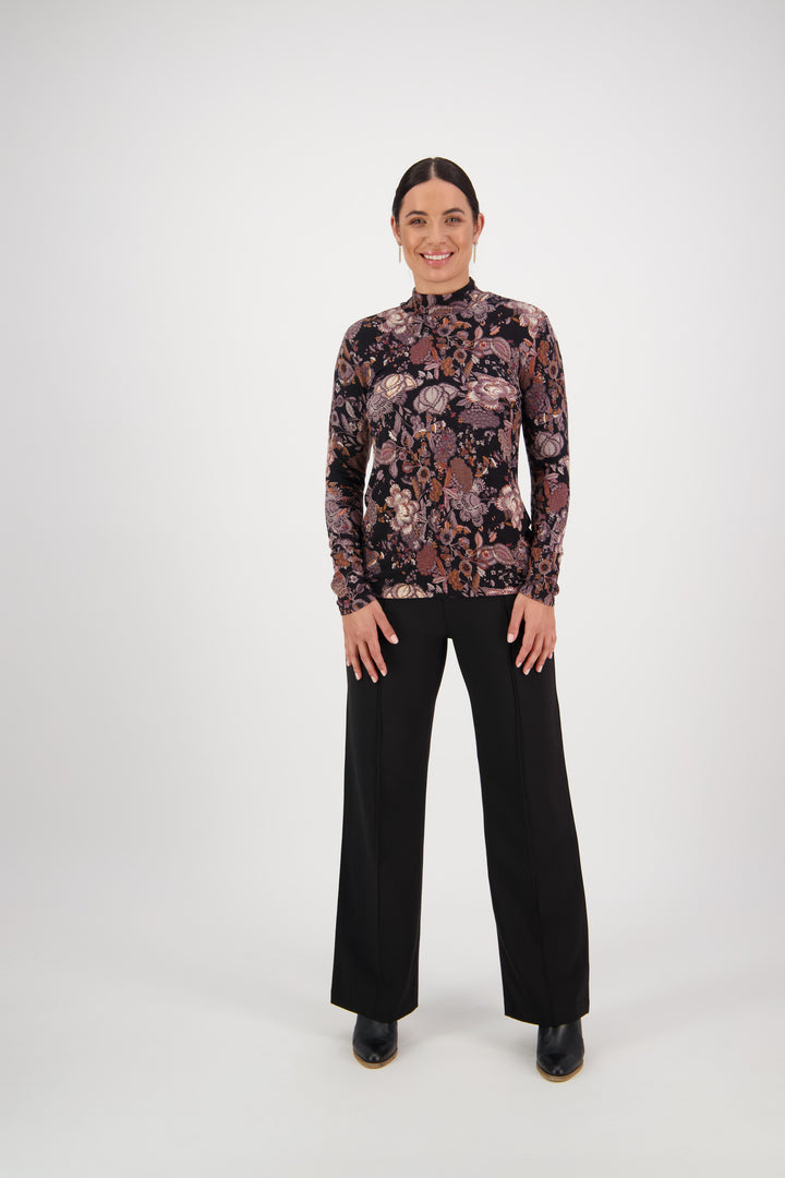 A person stands smiling against a plain white background, wearing the Vassalli Foundation Top - Vintage, paired with black wide-leg pants and black shoes.