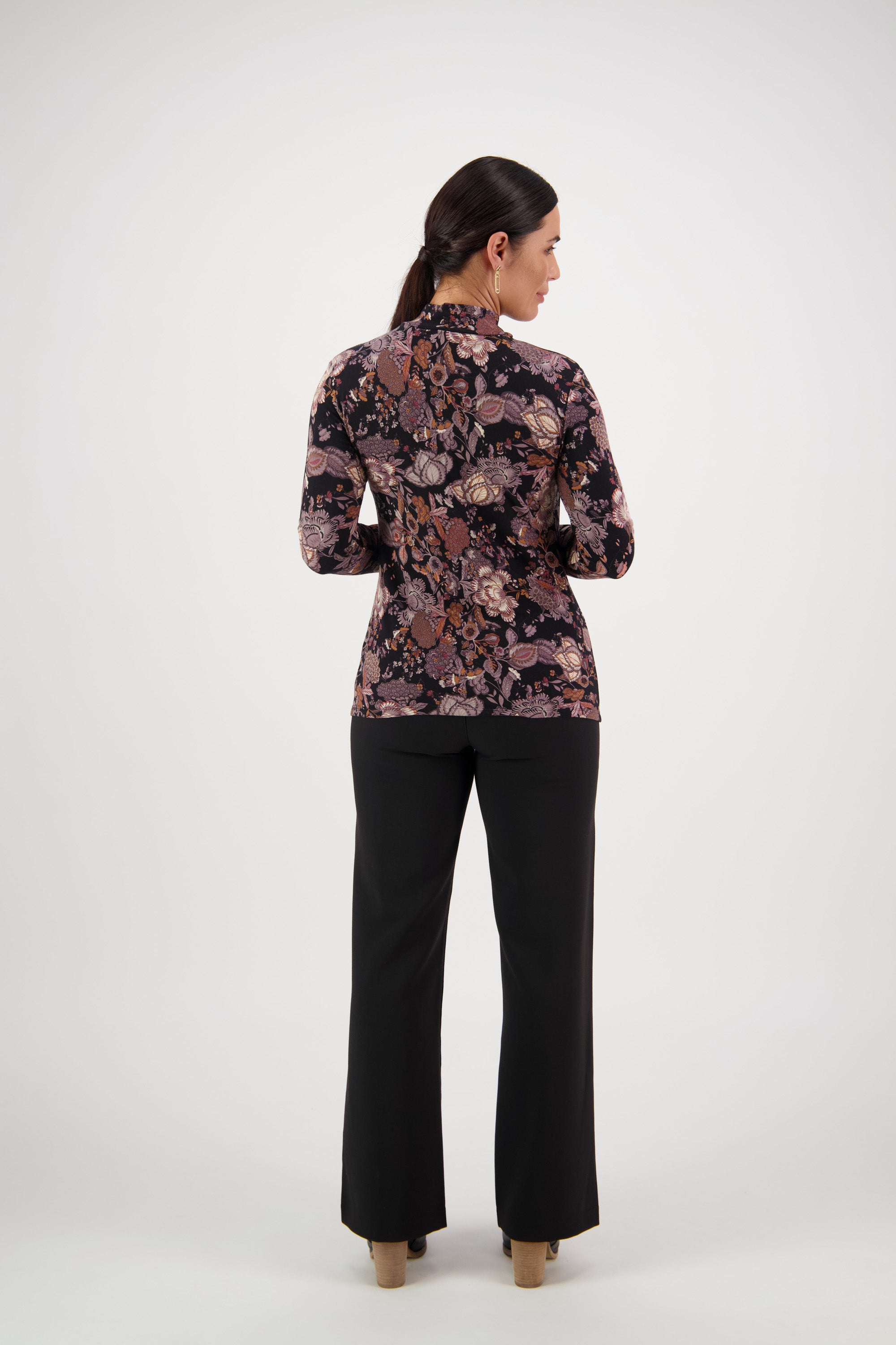 A woman with tied-back dark hair faces away, wearing the Vassalli Foundation Top - Vintage in black, paired with black pants and tan heeled shoes, set against a plain white background.