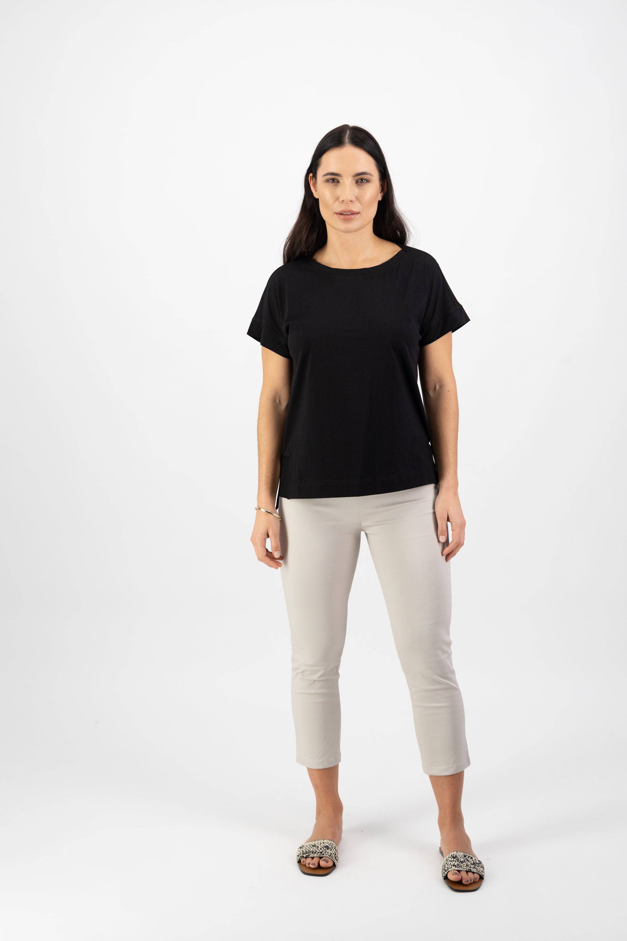 A woman with long dark hair stands facing forward, wearing the Vassalli Thea Top in black, light beige cropped pants, and leopard-print slip-on shoes against a plain white background.