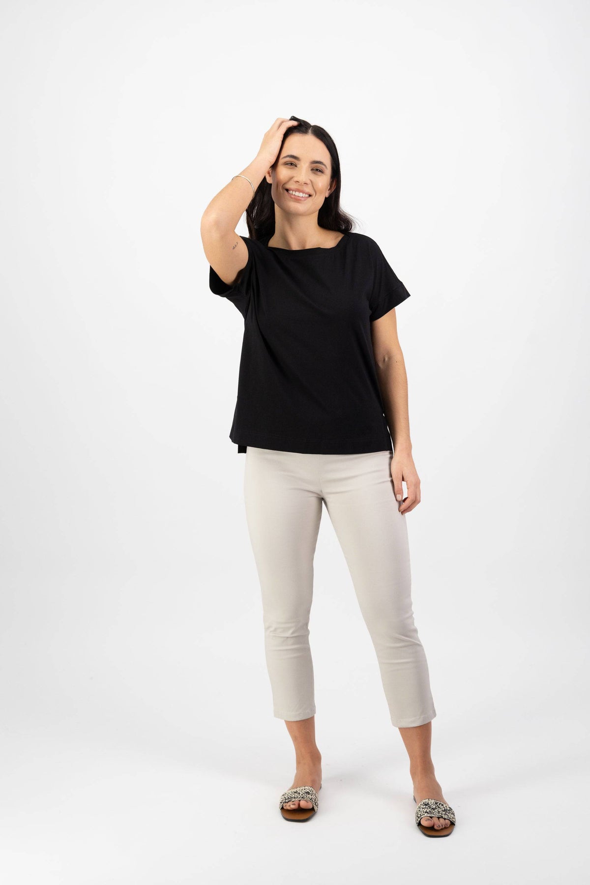A woman smiles against a white background, looking relaxed and cheerful as she wears the Vassalli Thea Top in black, paired with light cropped pants and sandals. One hand touches her head.