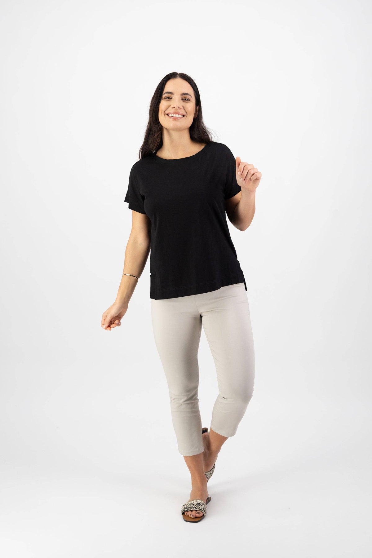 A woman with long dark hair smiles and poses against a white background, wearing the Vassalli Thea Top in black paired with light cropped pants and sandals.