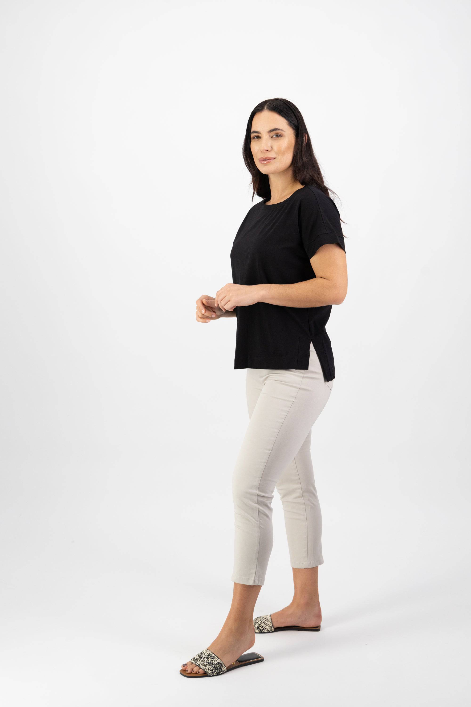 A woman with long dark hair stands sideways, facing the camera in a Vassalli Thea Top - Black, paired with light beige cropped pants and patterned slide sandals against a plain white background.